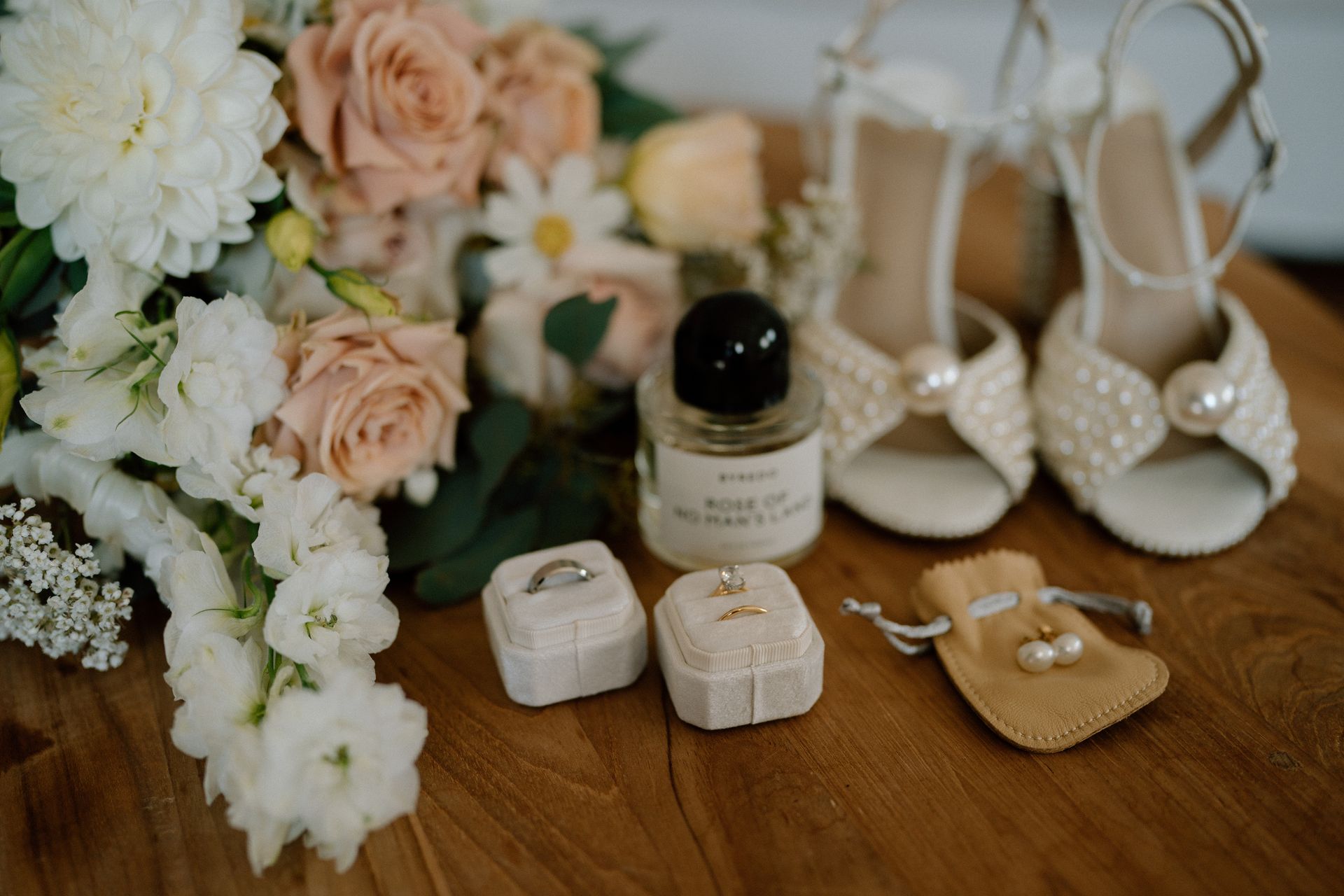 Wedding accessories on a wooden table with flowers, perfume, pearl heels, rings, and tan sandals