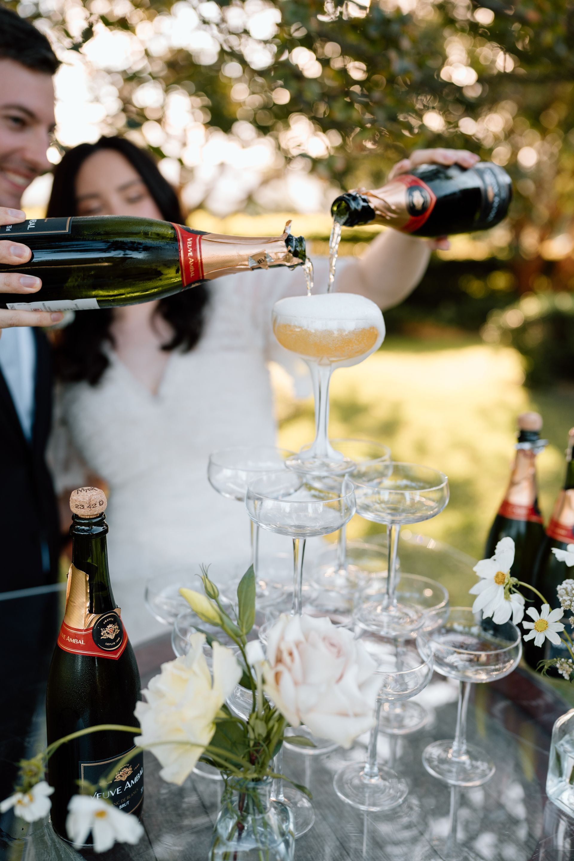 Champagne poured into stacked glasses at an outdoor celebration table with flowers and bottles