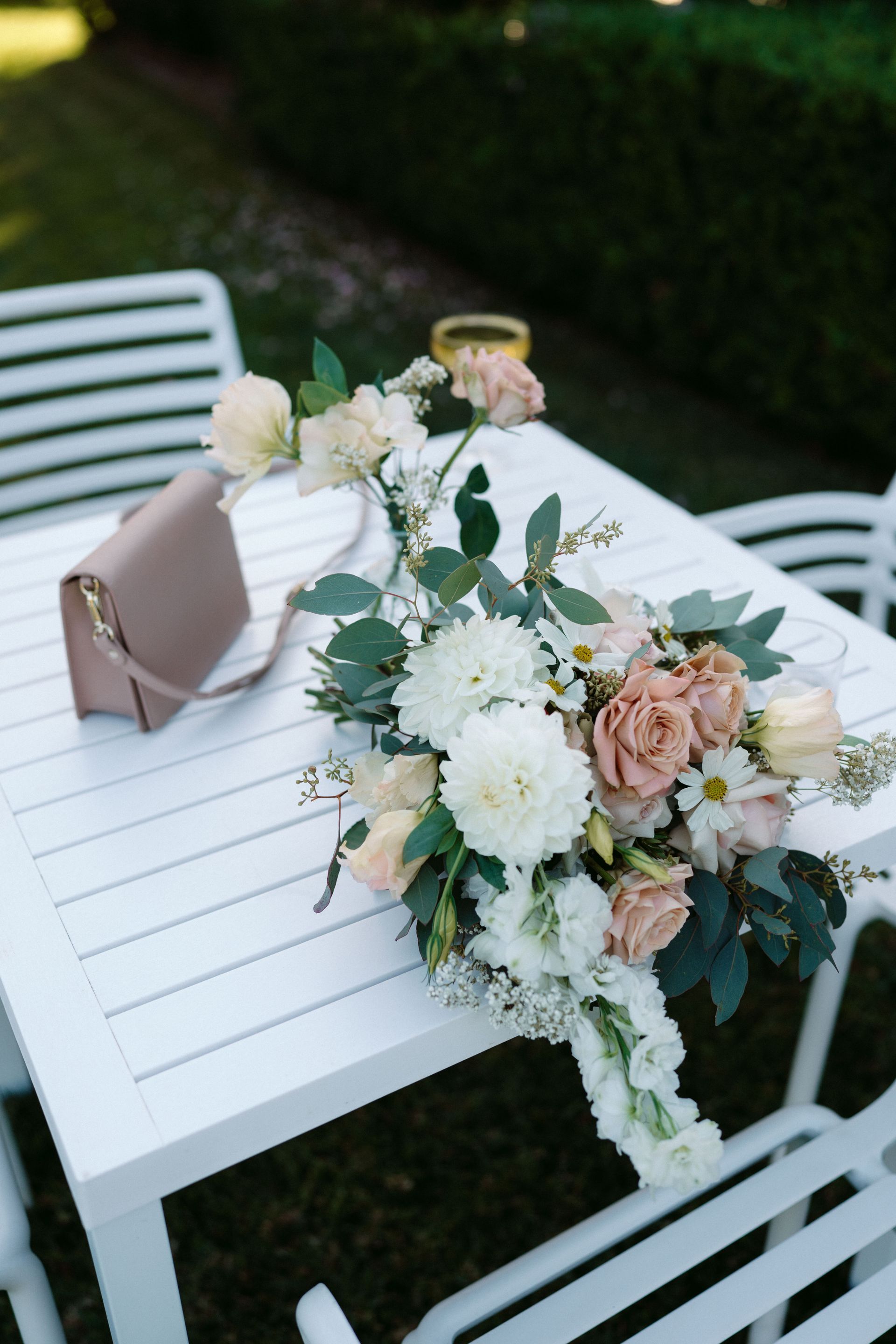 White table with blush and white floral bouquet and beige clutch on a garden patio