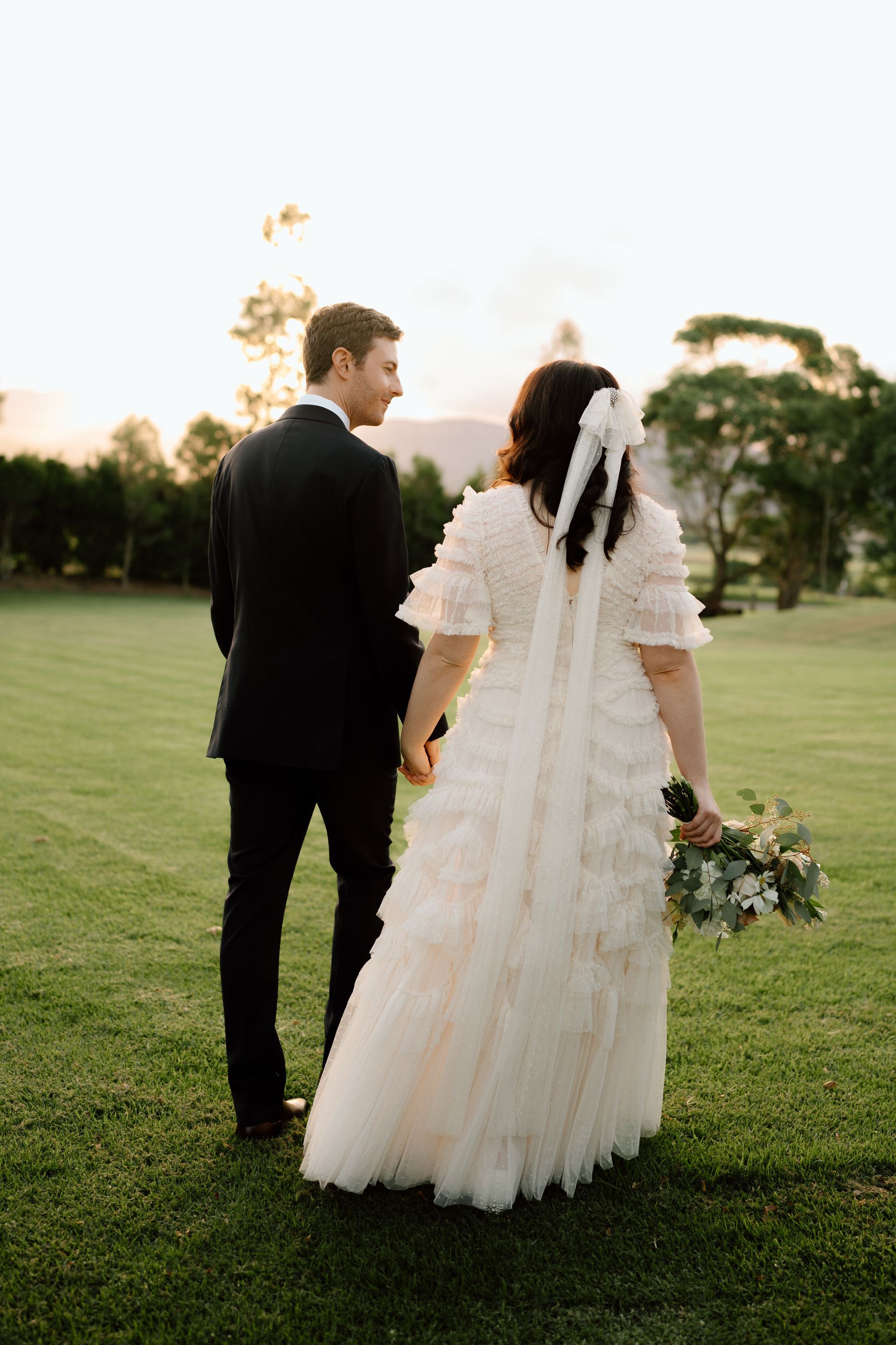 Couple in formal attire holding hands on a grassy field at sunset, the bride carrying a bouquet.