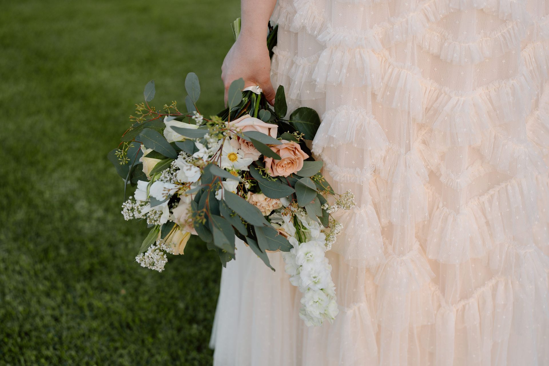 Hand holding a pastel bouquet beside a cream ruffled dress on green grass
