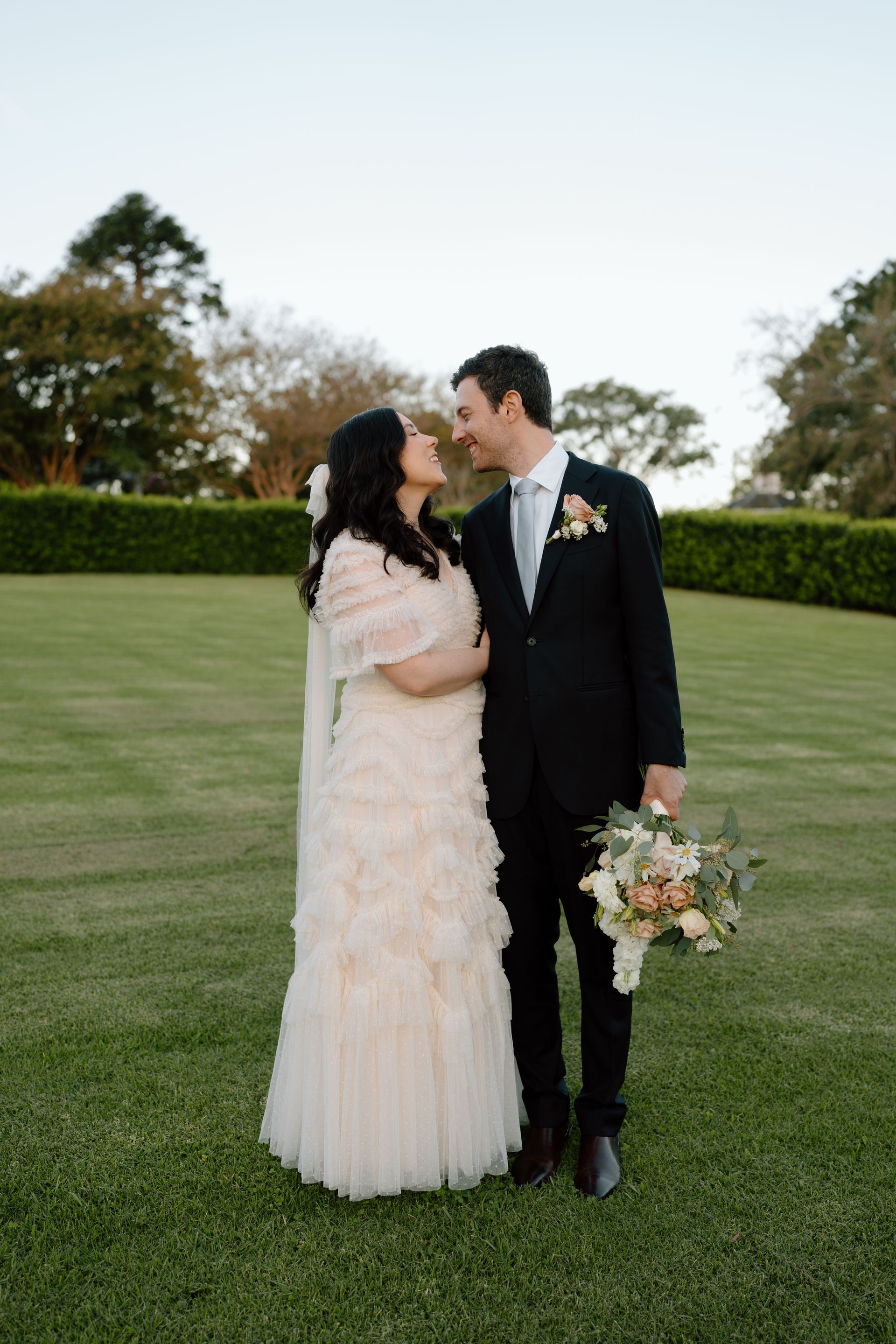 Couple in formal attire embracing on a lawn, holding a bouquet at an outdoor wedding venue.