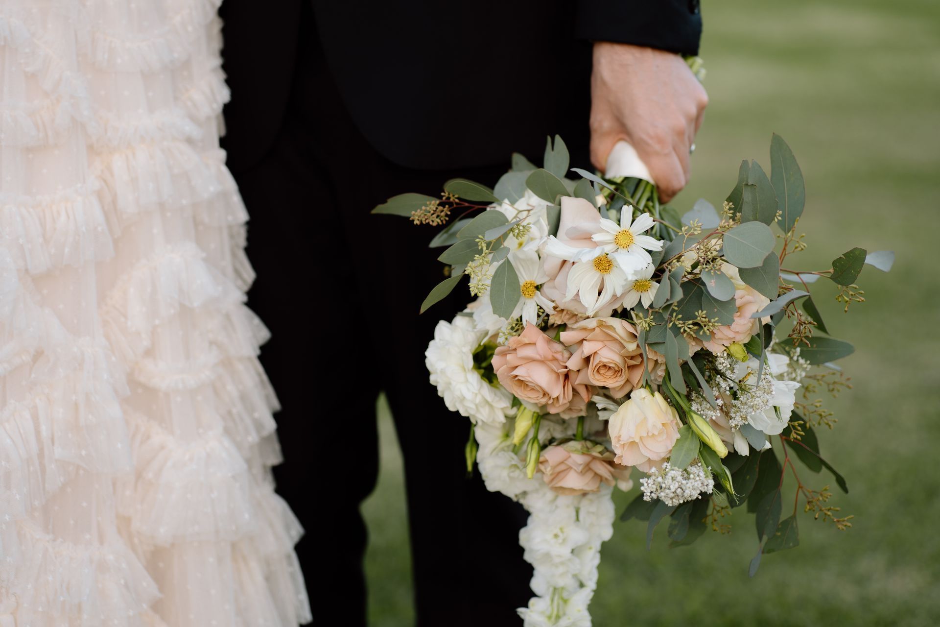 Wedding bouquet held by a couple, with white, blush, and green flowers beside a cream dress and black suit