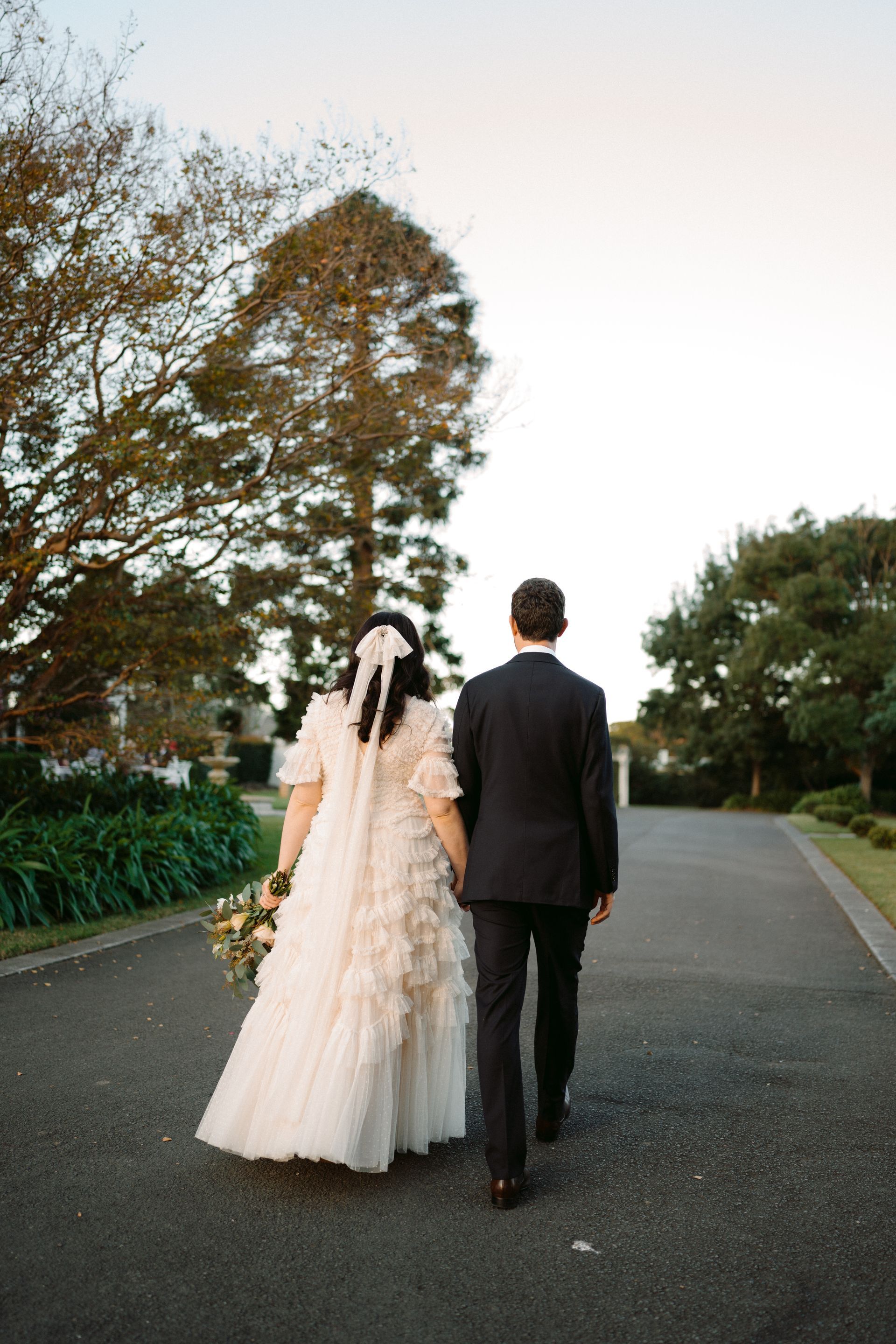 Bride and groom walking away down a quiet road at dusk, bride in a white gown and groom in a black suit