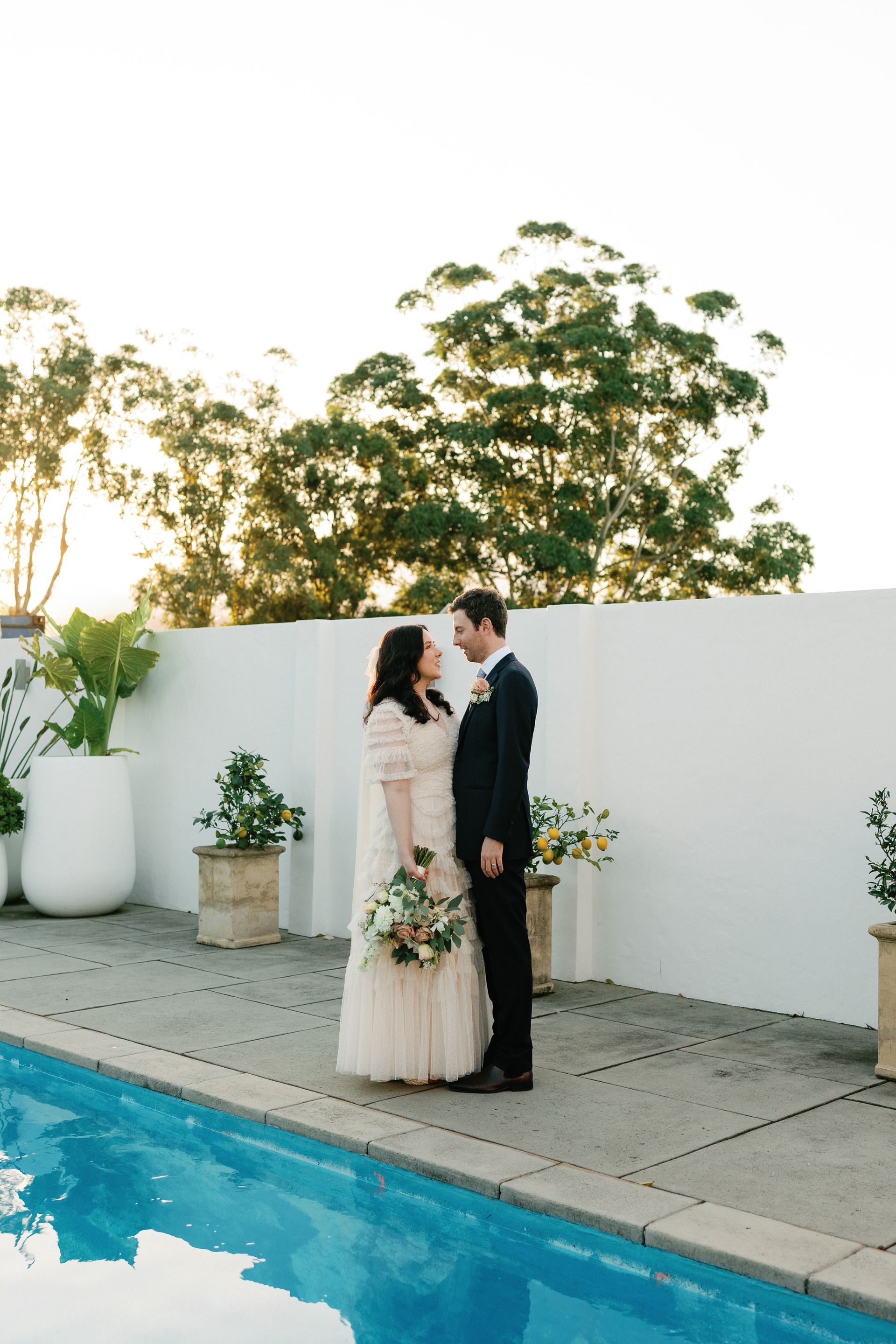 Couple in wedding attire embracing beside a pool at sunset, with potted plants and a white wall backdrop