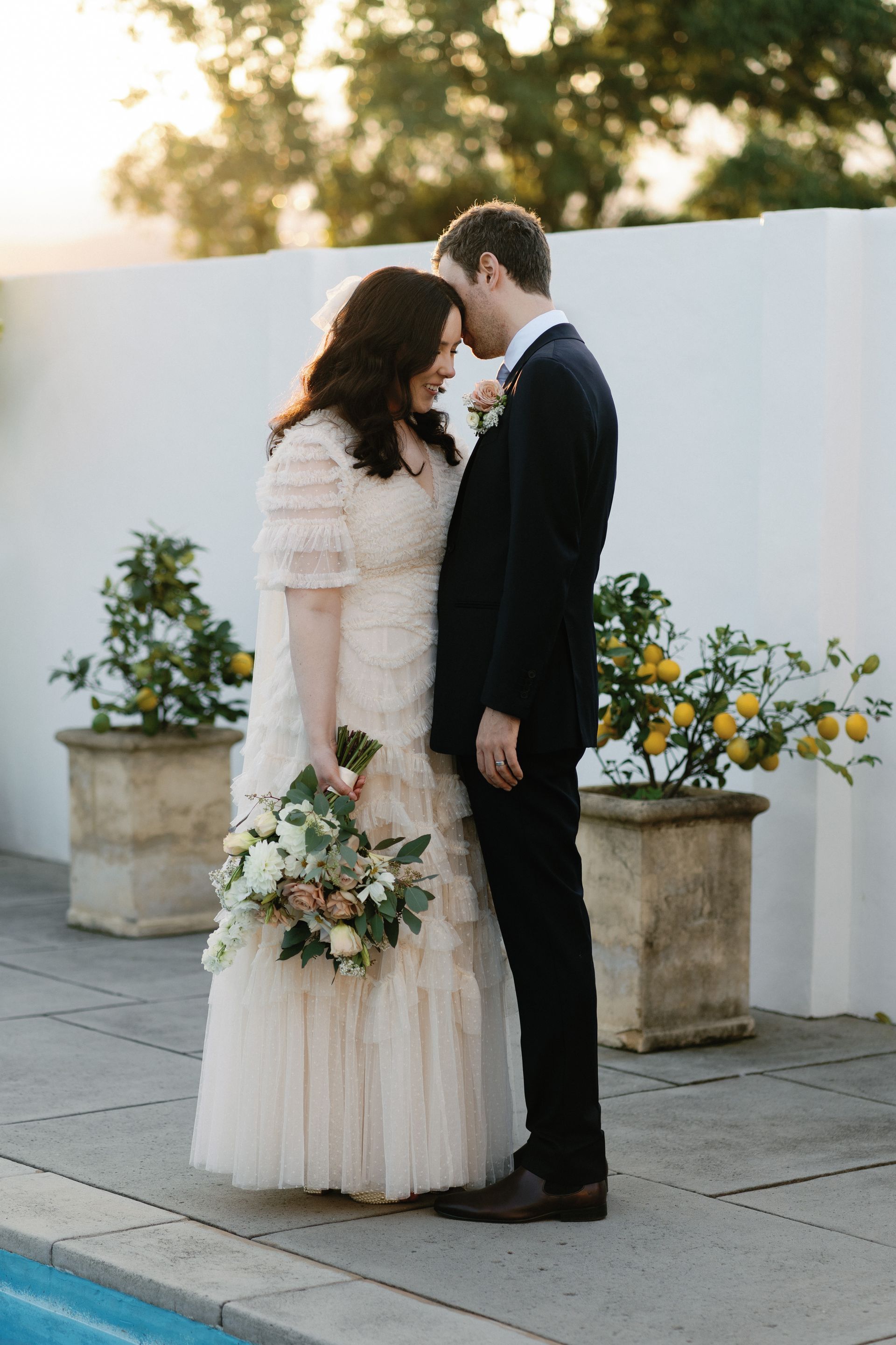A bride and groom embrace outdoors by potted flowers at sunset.