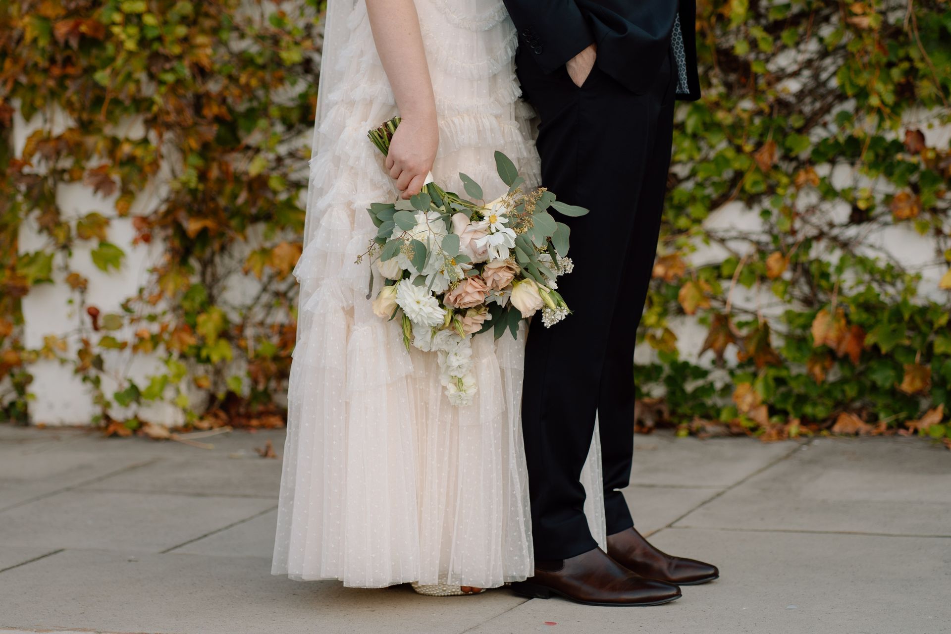 Bride and groom standing together, holding a pastel bouquet against a leafy wall