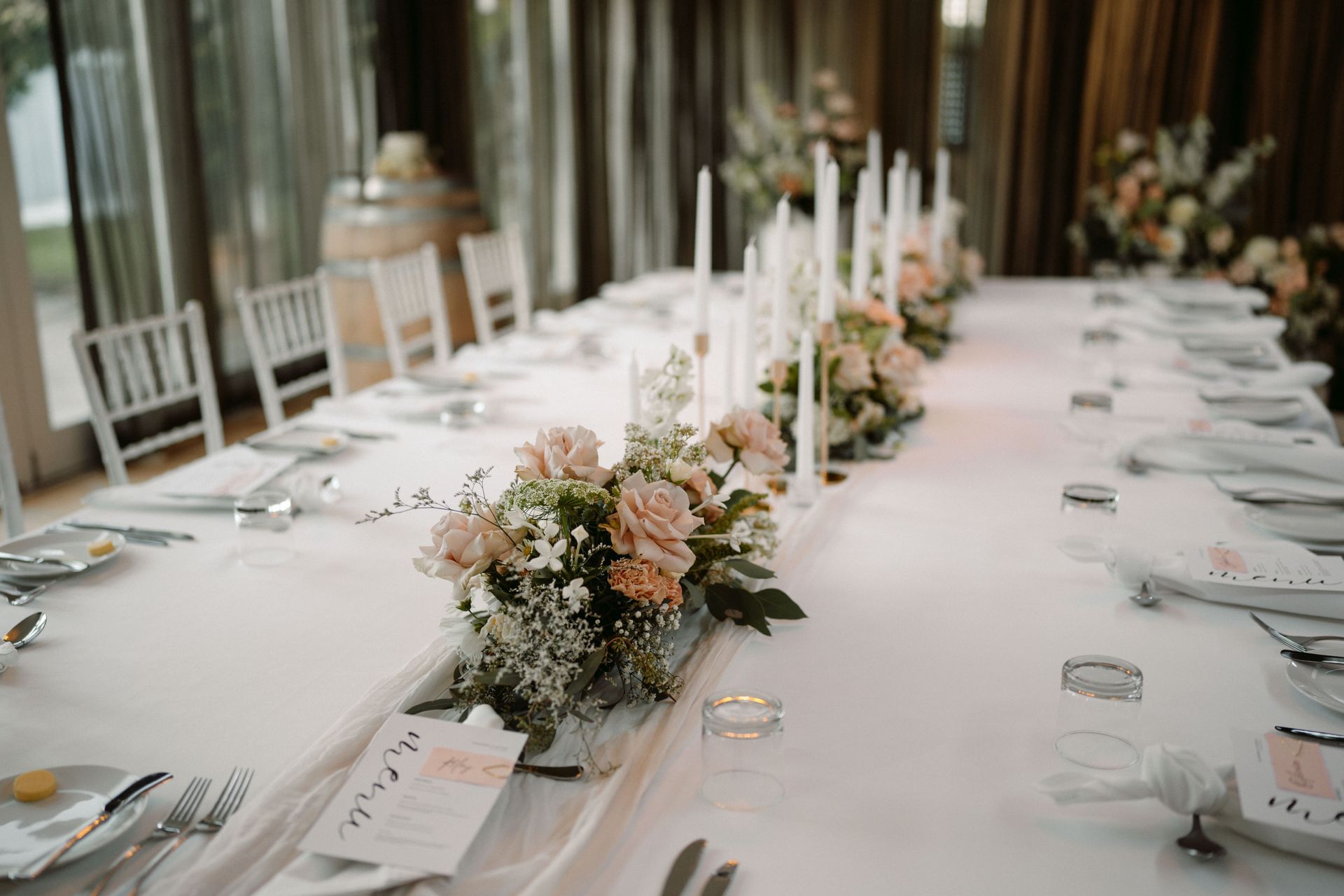 Elegant long banquet table with white linens, candles, and blush floral centerpieces in a bright reception room
