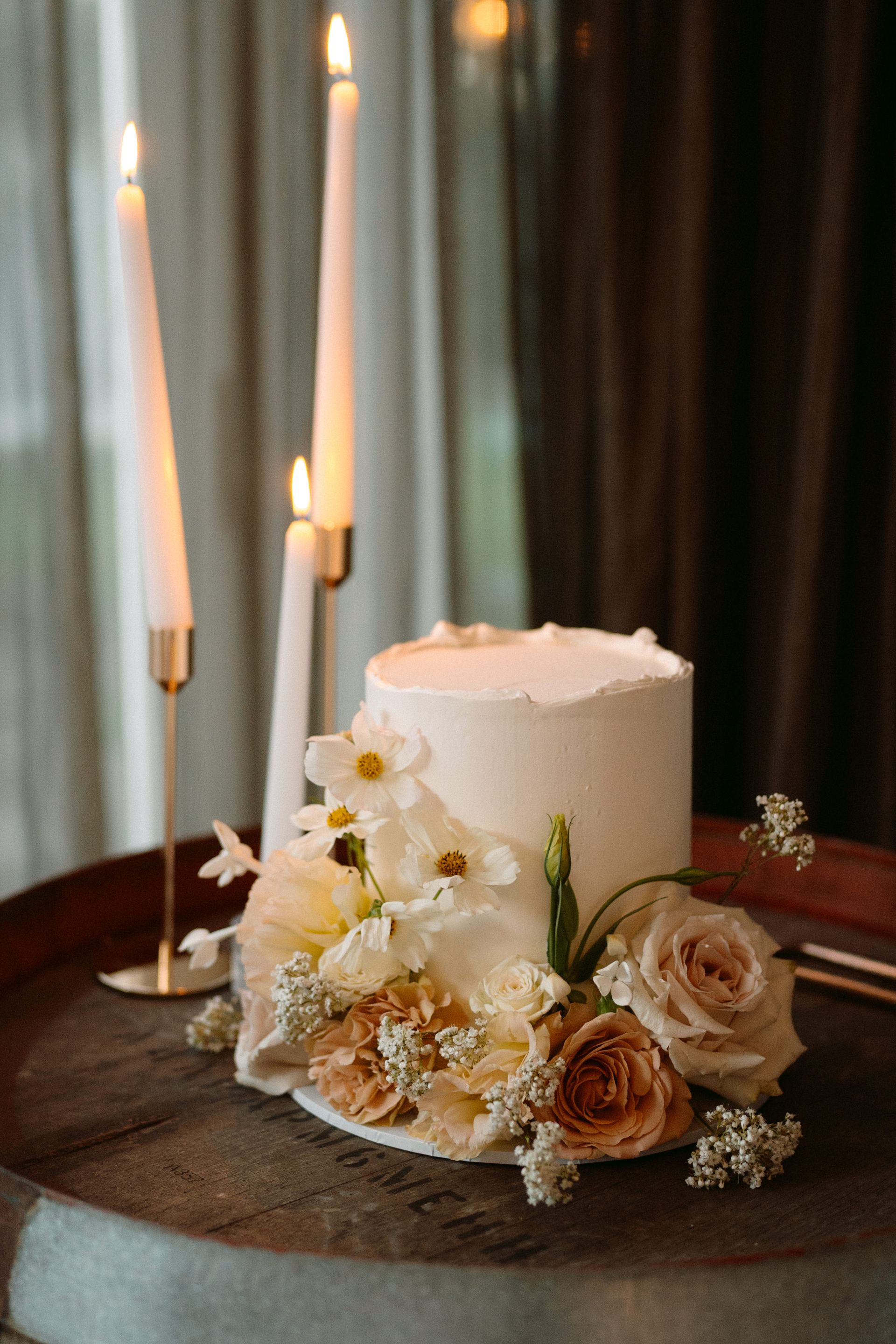 White cake with floral decorations and lit candles on a table by a window.