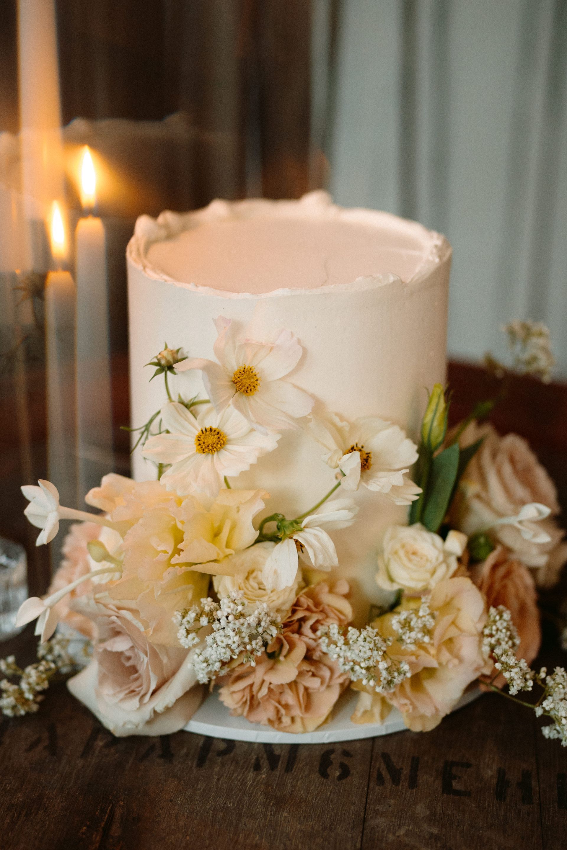 White floral wedding cake with cream and blush flowers on a table beside candles