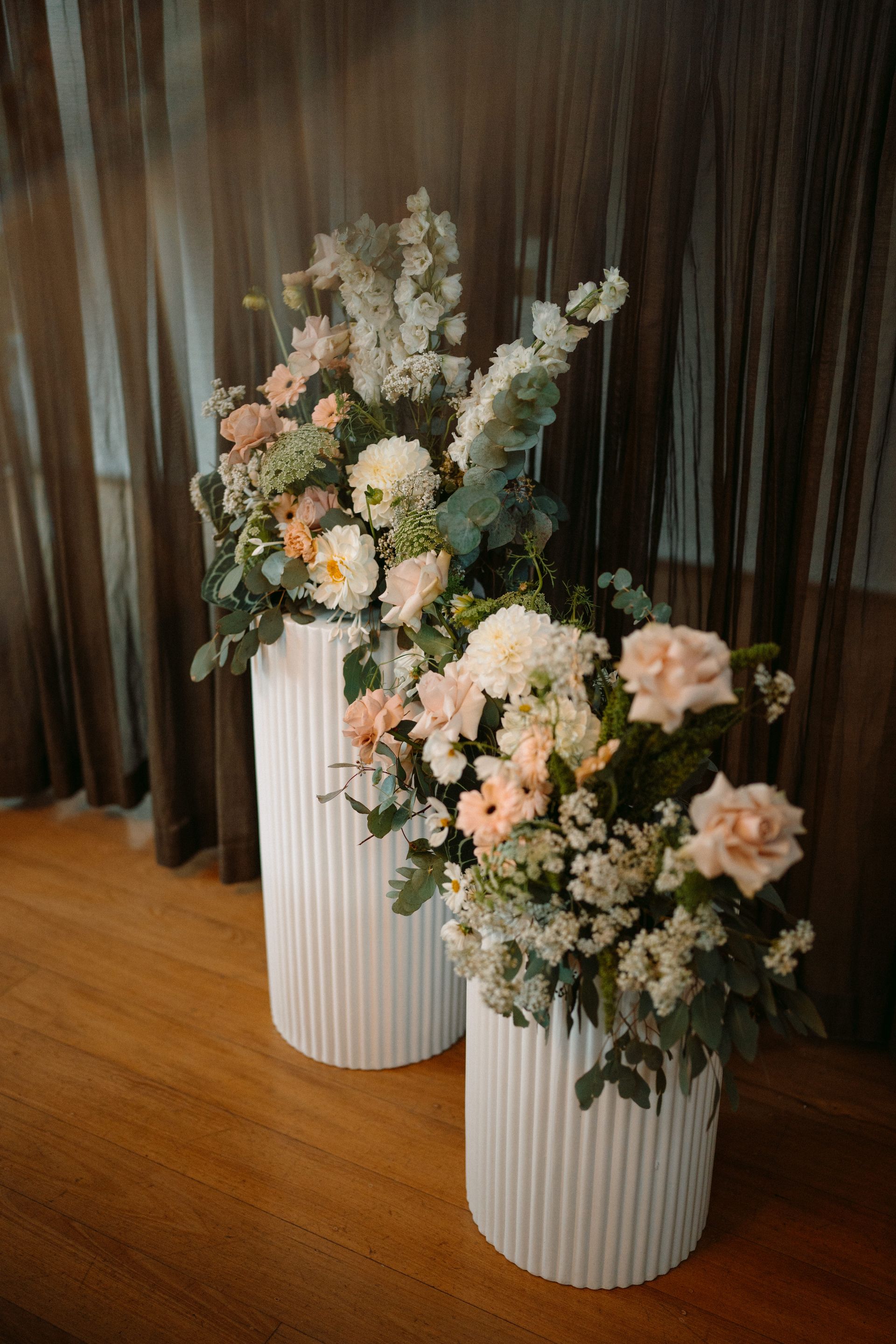Two white ribbed vases with peach and white floral arrangements on a wooden floor by dark curtains