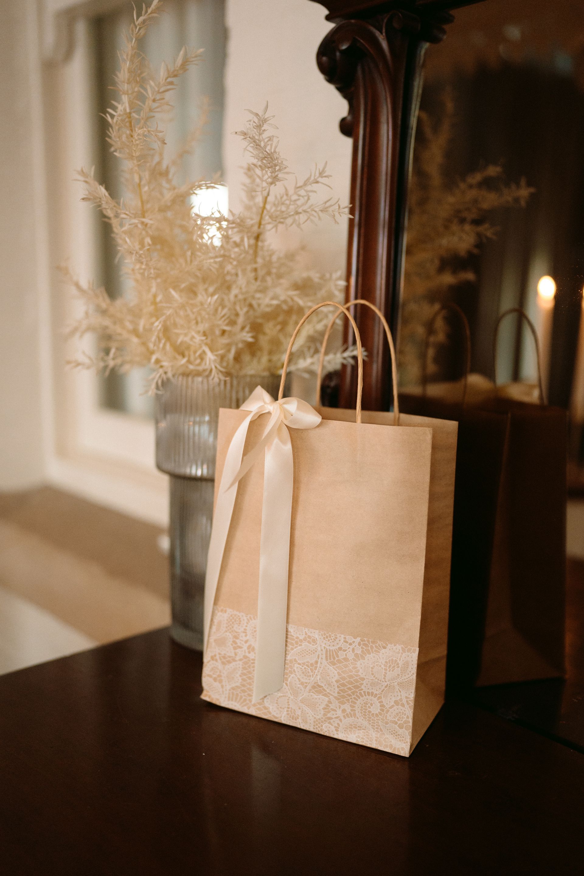 Kraft gift bag with cream ribbon on a dark table beside a vase of dried flowers and a mirror