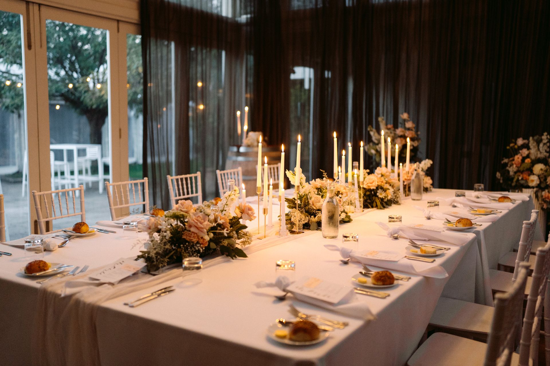 Elegant candlelit banquet table set in a glass-walled dining room with white linens and floral centerpieces