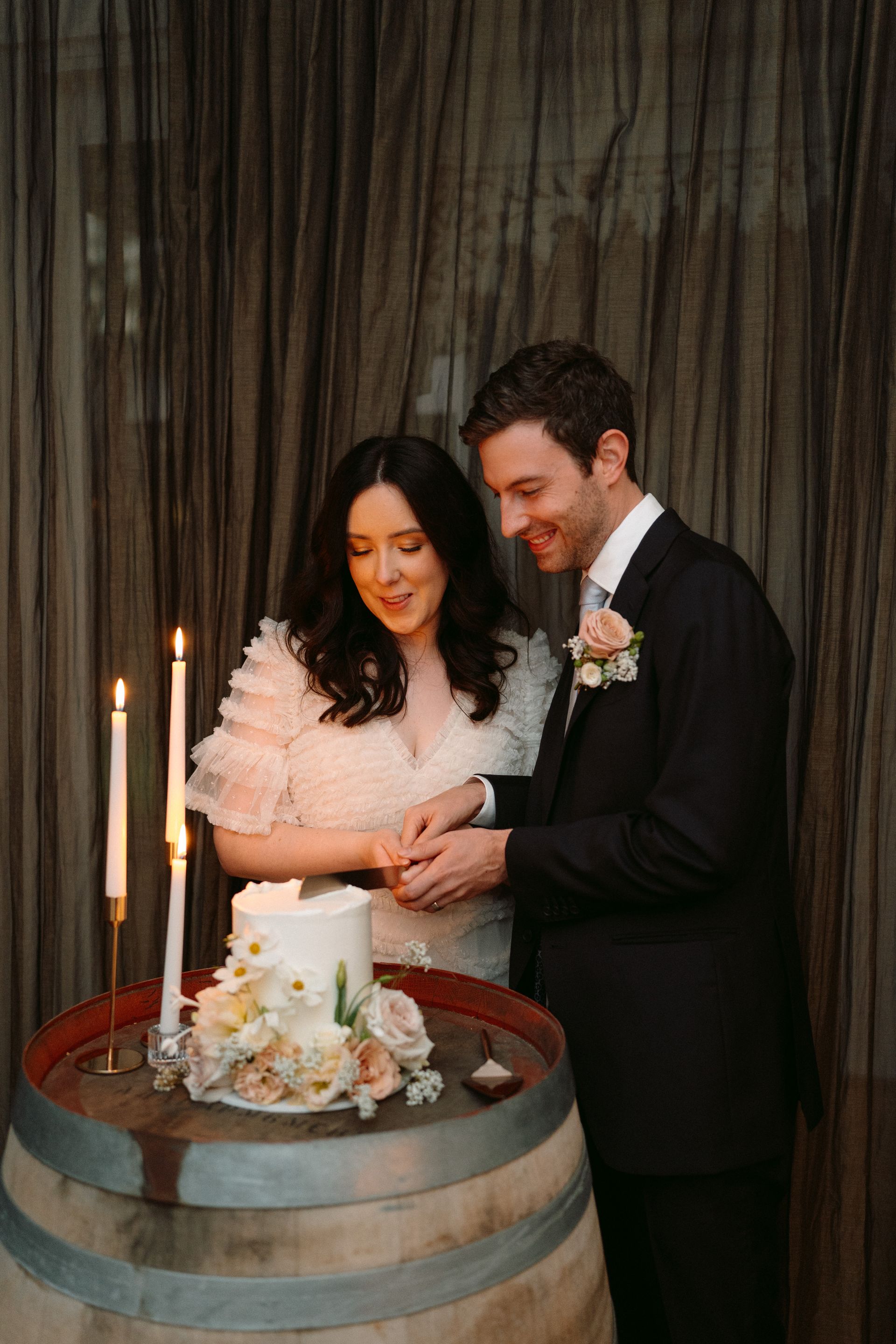 Couple cutting a small white wedding cake beside lit candles in a dimly lit room