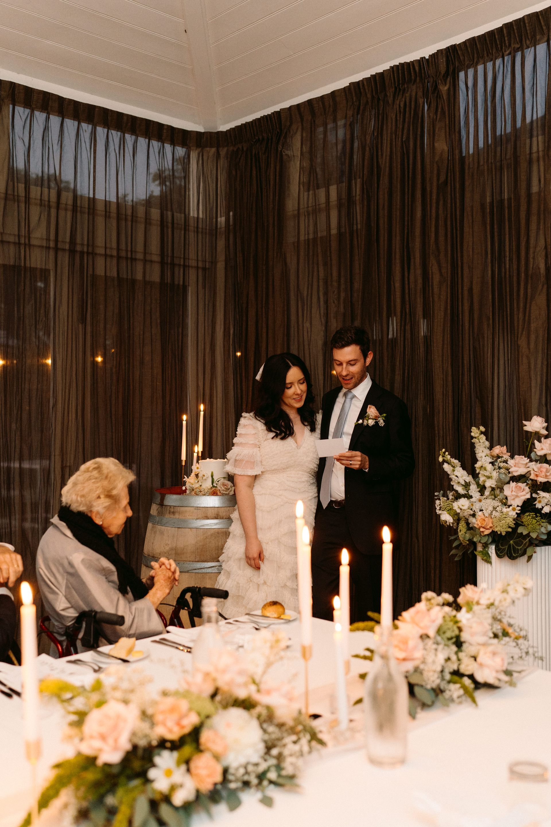 Wedding couple at a candlelit table, with flowers and sheer curtains in the background