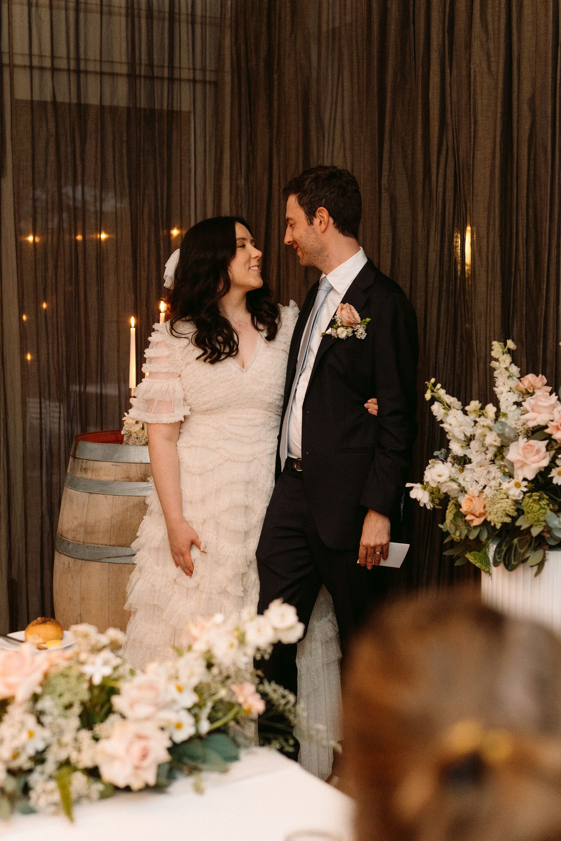 Couple in formal attire smiling at each other beside a floral arrangement at a wedding reception