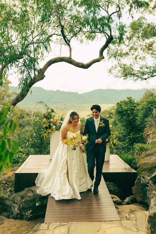 Bride and groom walking on a wooden bridge in a lush garden, holding flowers under a leafy arch.