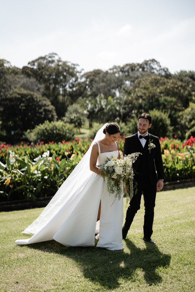 Bride in white gown and groom in dark suit standing on grass in a garden, sharing a bouquet
