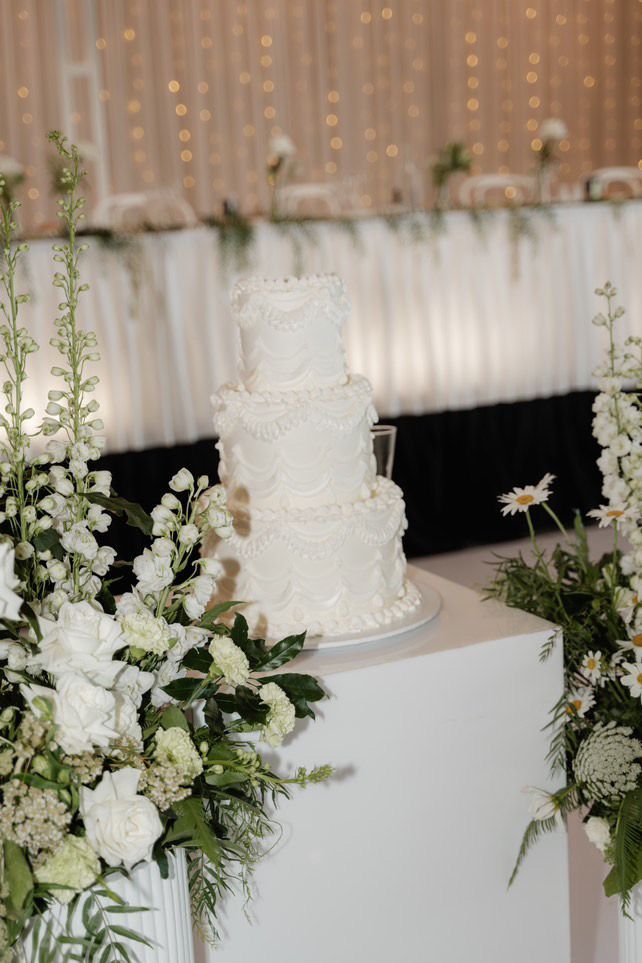 White tiered wedding cake with floral decorations on a pedestal, surrounded by white flower arrangements