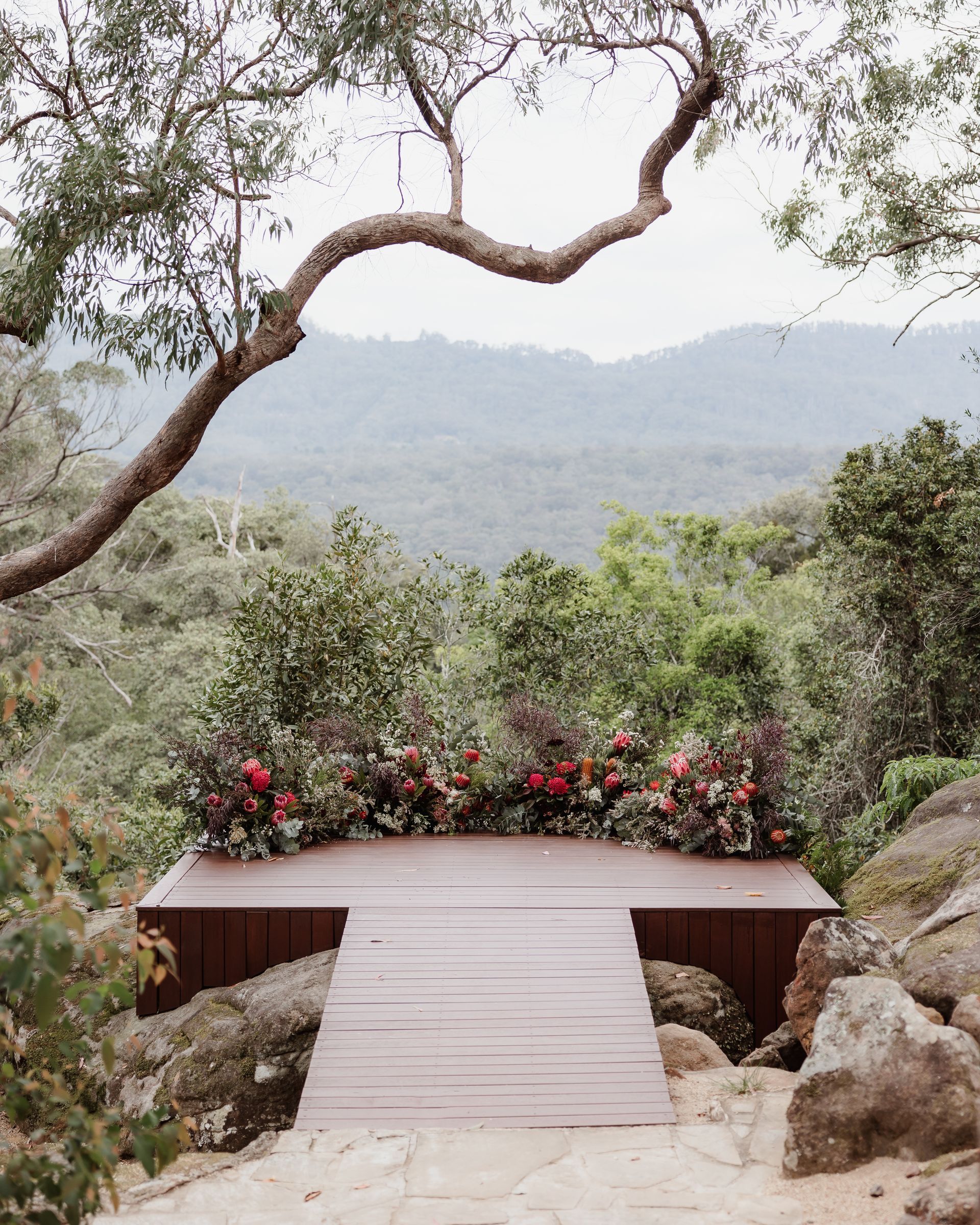 Wooden platform with floral arrangement overlooking a mountain range, under a tree with reaching branches.