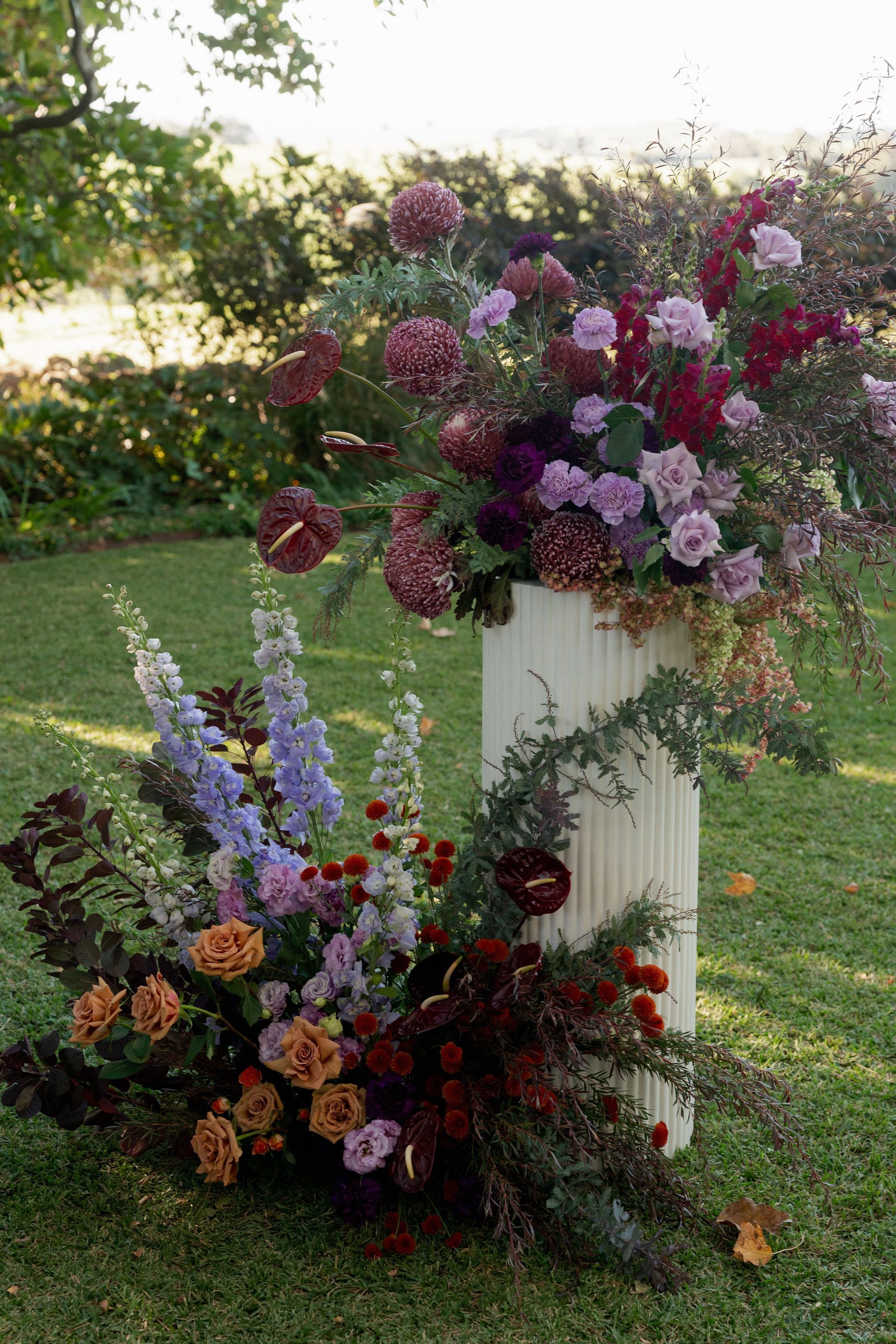 Floral arrangement on a white pedestal in a garden, with purple, pink, and orange flowers.