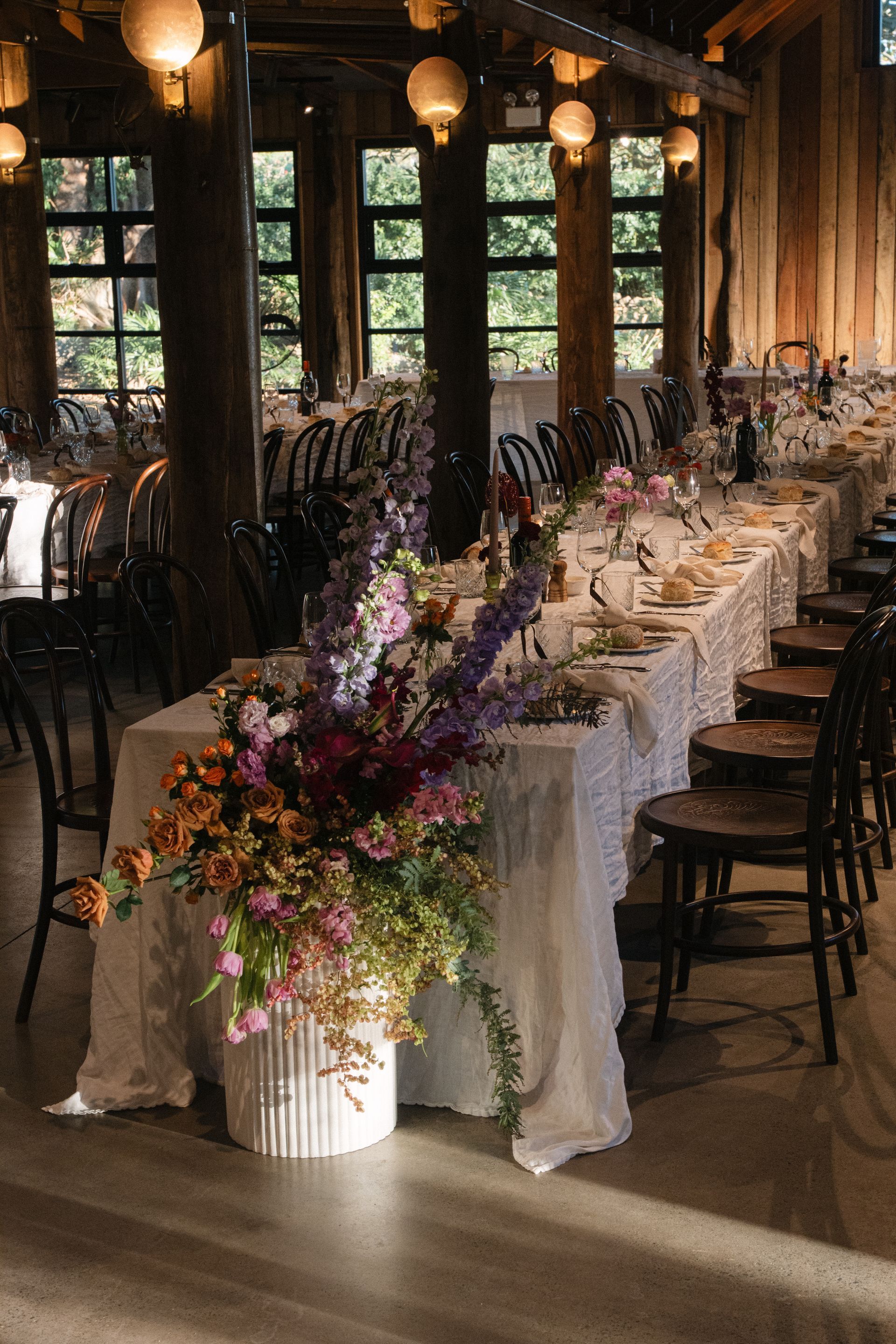 Long banquet table with white linens and purple floral arrangements in a rustic, wood-paneled room