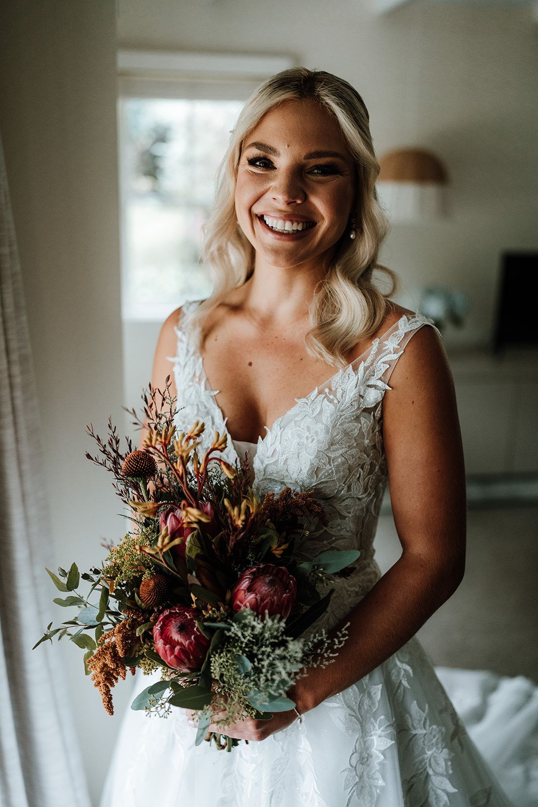 Smiling bride in a white lace wedding dress holding a colorful bouquet indoors