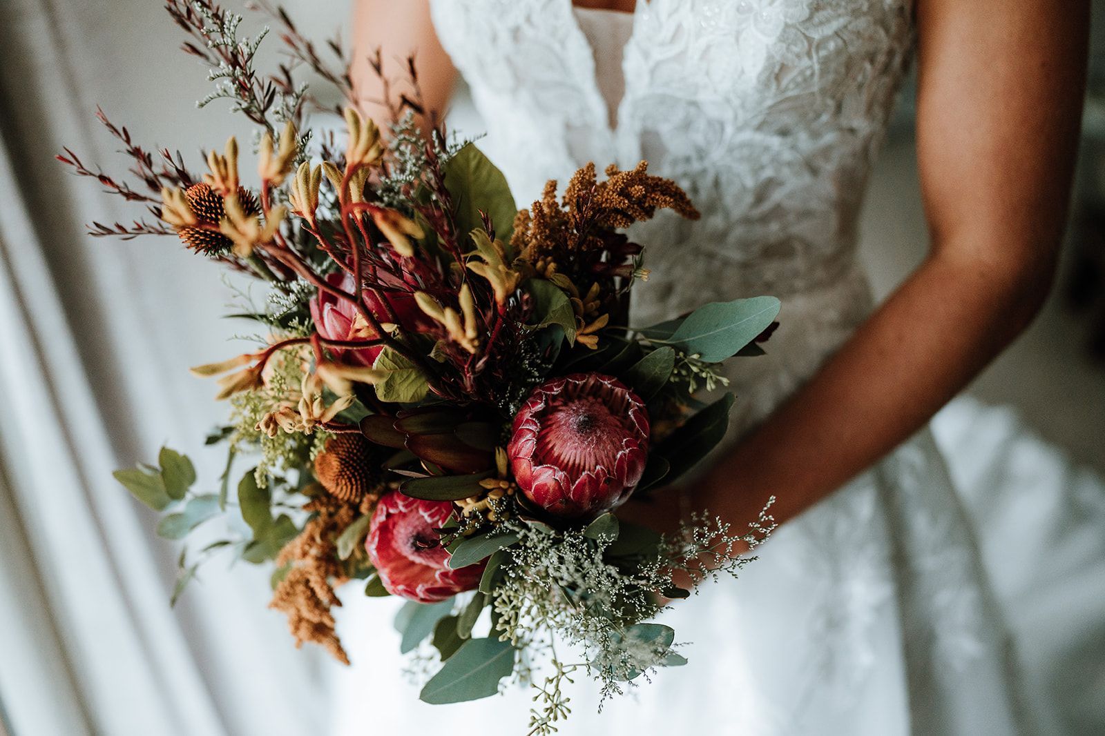 Bride holding a rustic bouquet of red, burgundy, and green flowers in a lace wedding dress