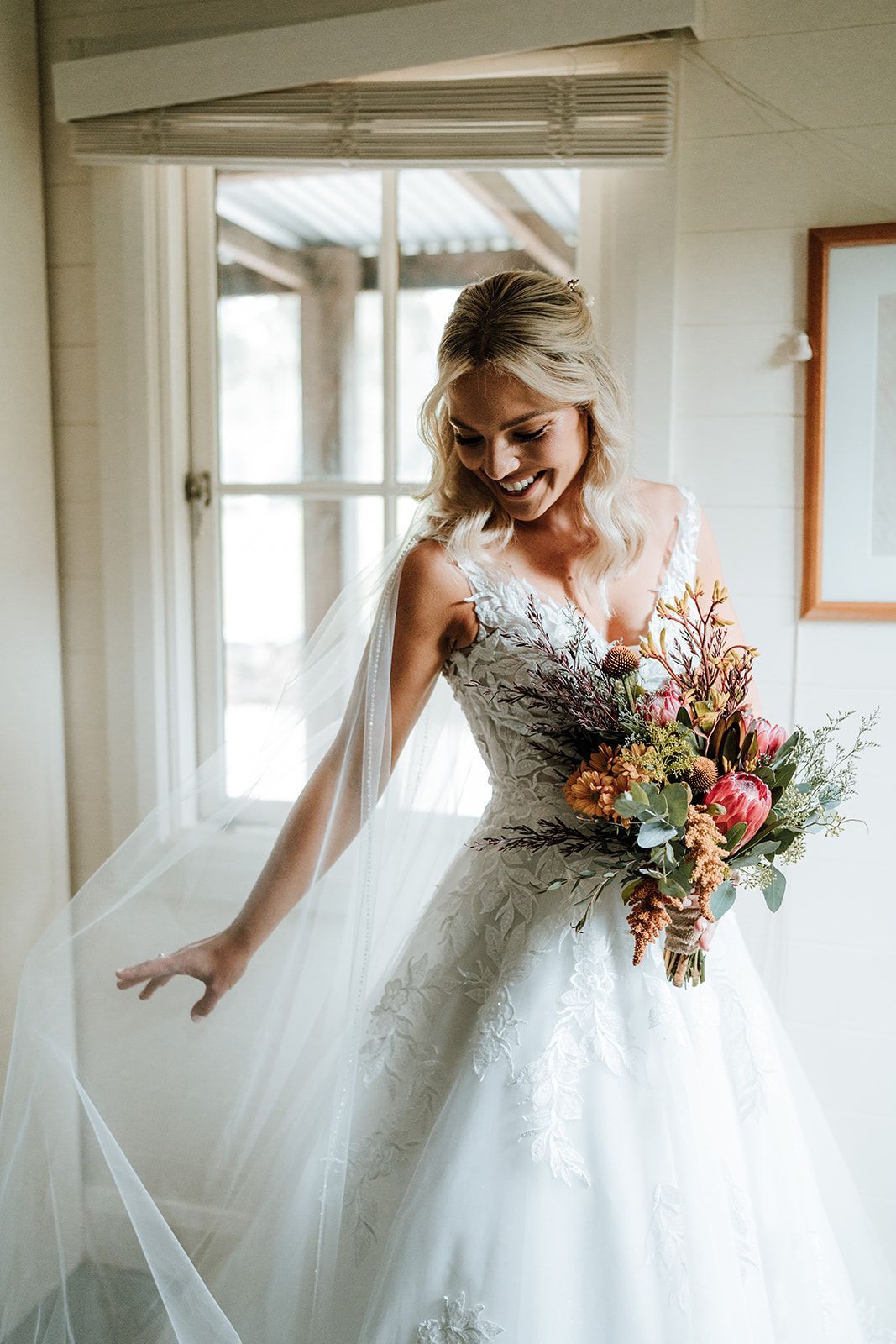 Bride in a white lace wedding gown holding a colorful bouquet, smiling by a doorway.