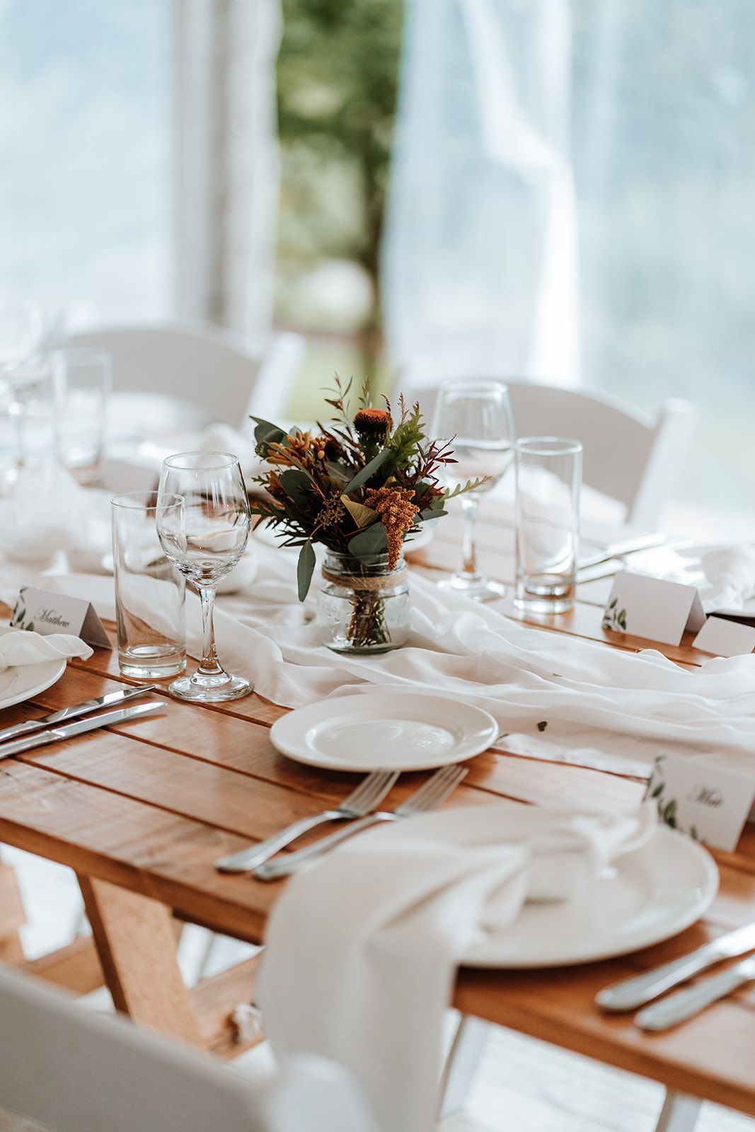 Elegant white-clothed dining table with plates, glasses, and a floral centerpiece by a bright window