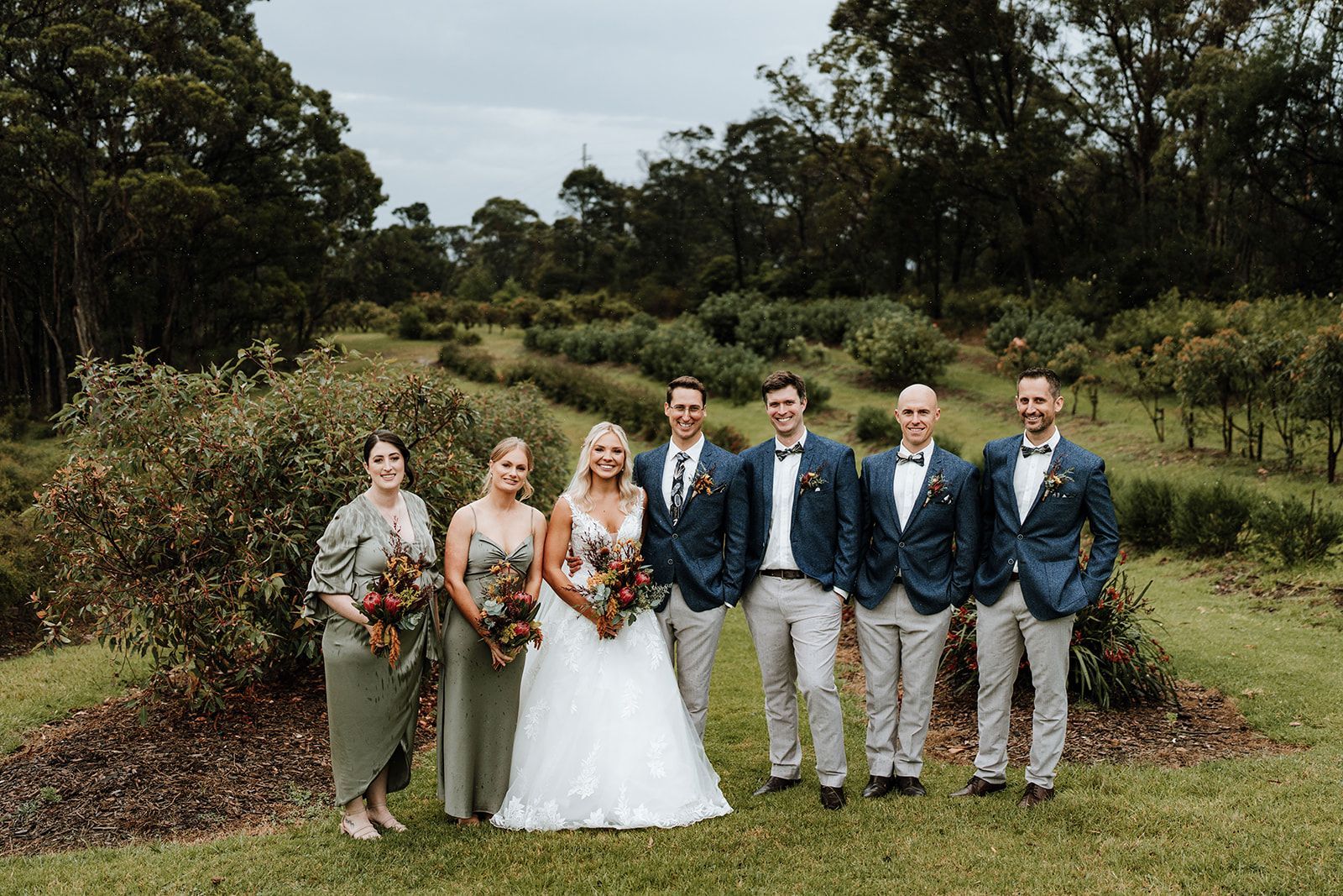 Wedding party posing outdoors in formal attire, with bride holding a bouquet in the center.