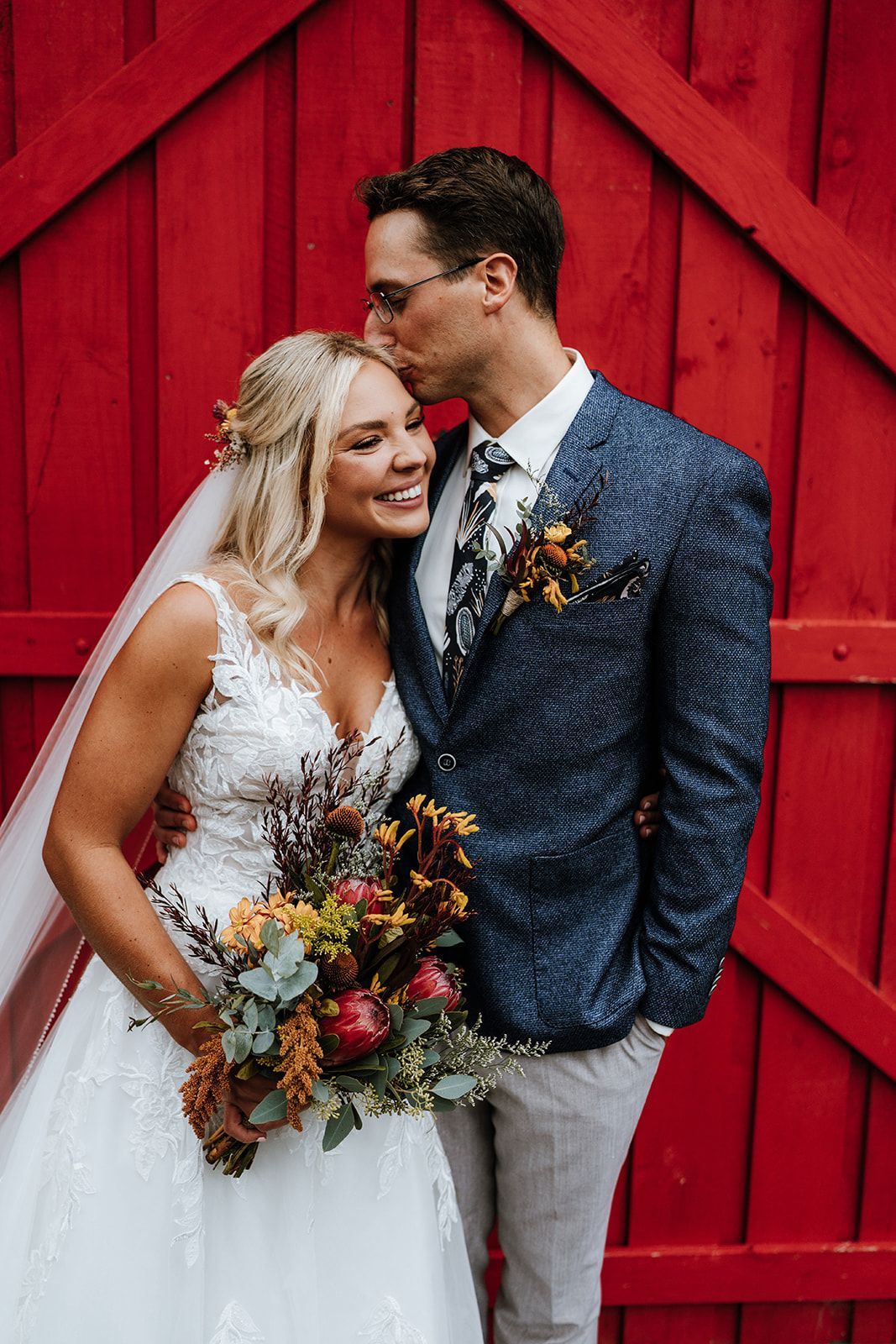 Wedding couple embracing in front of a bright red barn door, holding a colorful bouquet