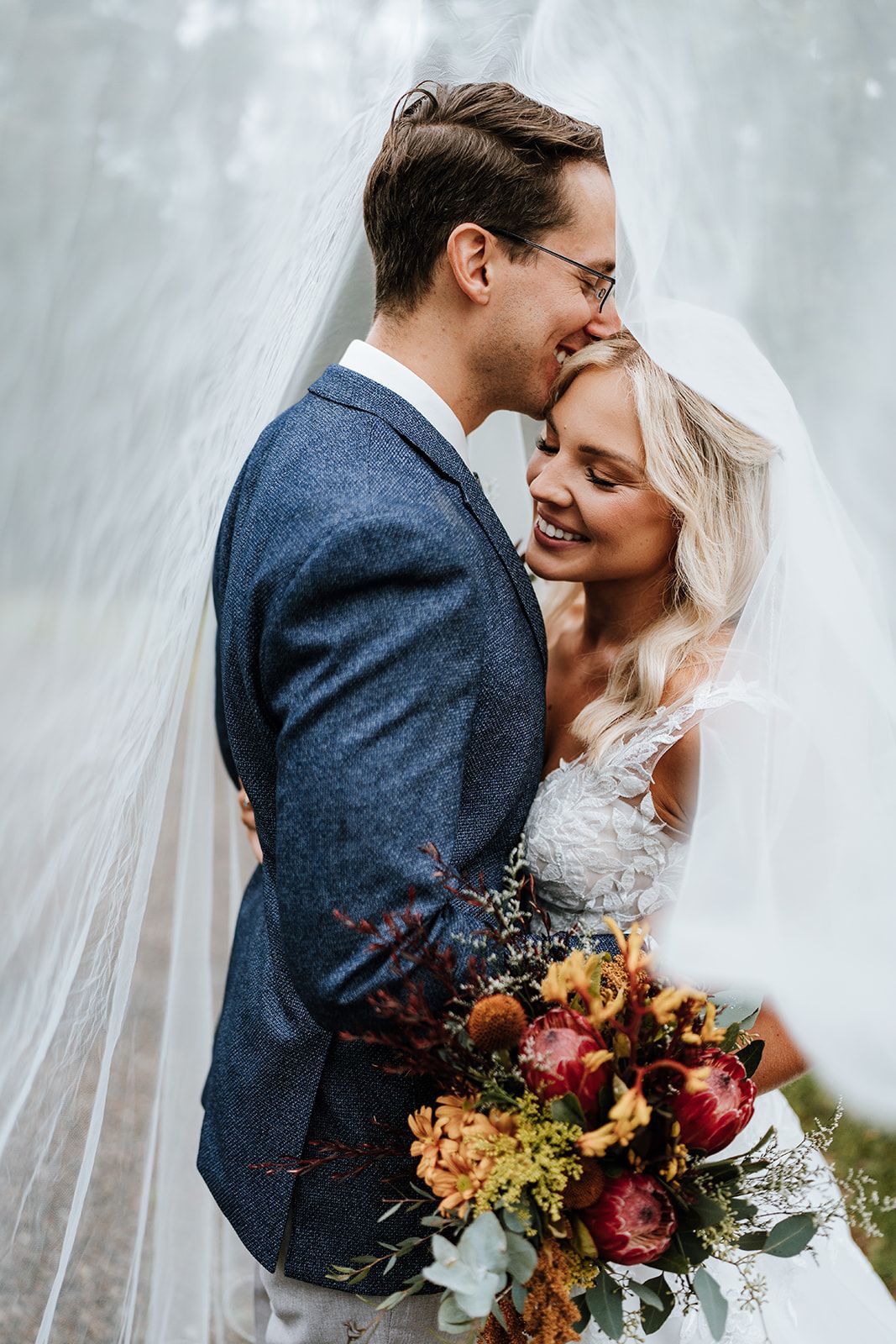 Bride and groom embracing beside a waterfall, smiling with a colorful bouquet and veil flowing