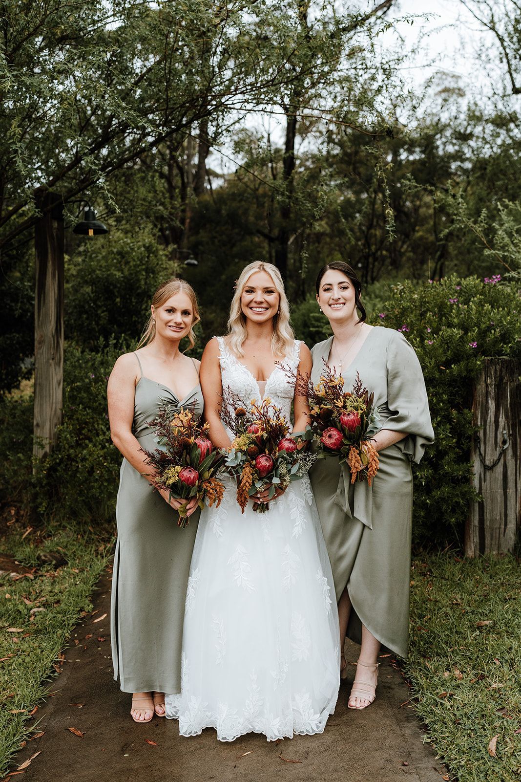 Three women in formal dresses holding bouquets outdoors on a grassy path.