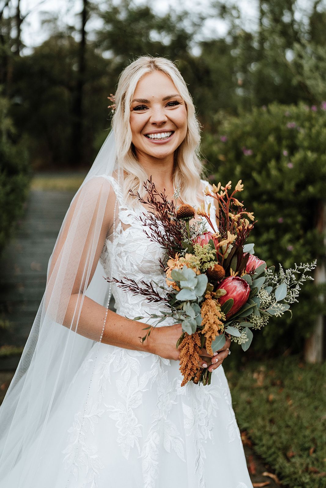 Bride in a white lace gown and veil, smiling outdoors while holding a bouquet of flowers.