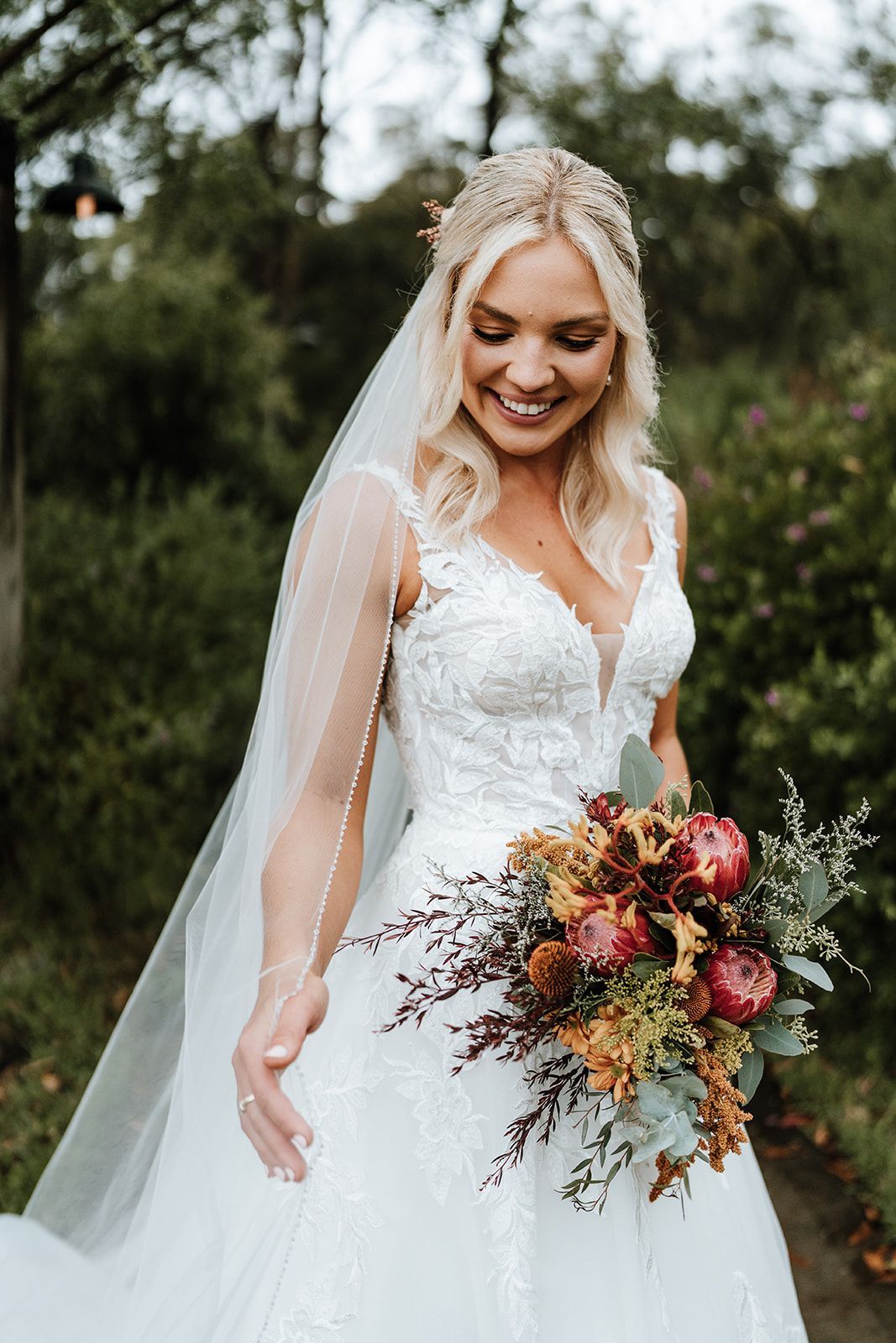 Bride in a white lace gown holding a colorful bouquet outdoors, smiling and looking down