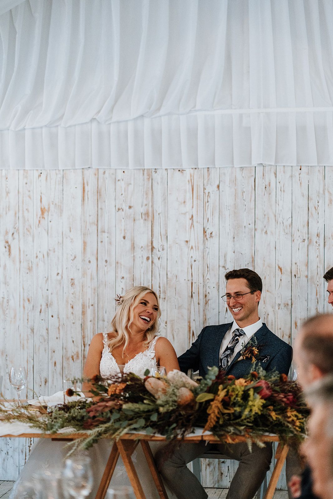 Wedding couple seated at a floral-decorated table, smiling under white drapery