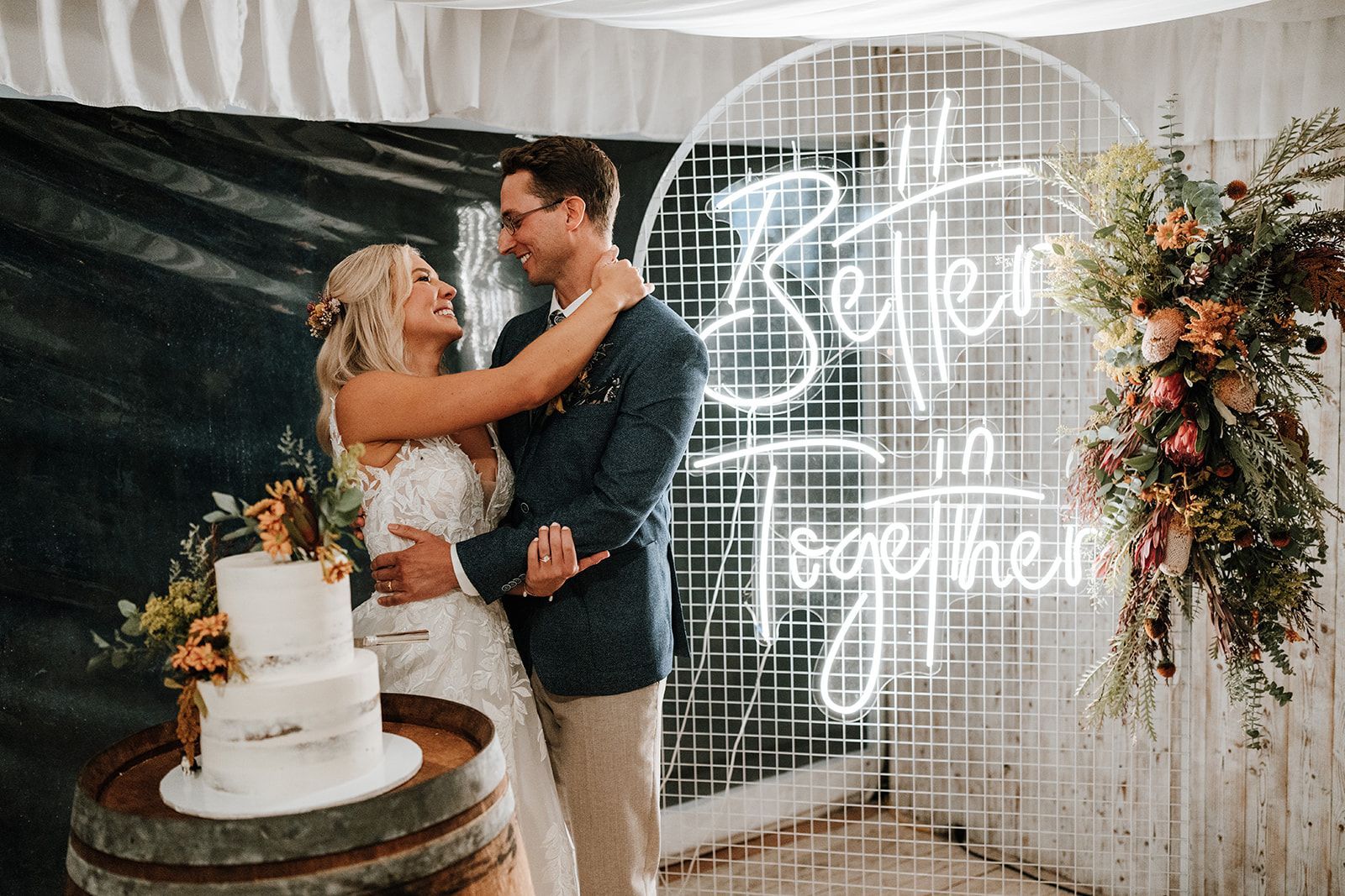 Couple embracing at a wedding reception beside a cake and glowing “Better Together” sign