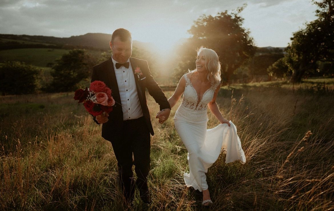Bride and groom walking through a sunlit field, holding hands and a bouquet