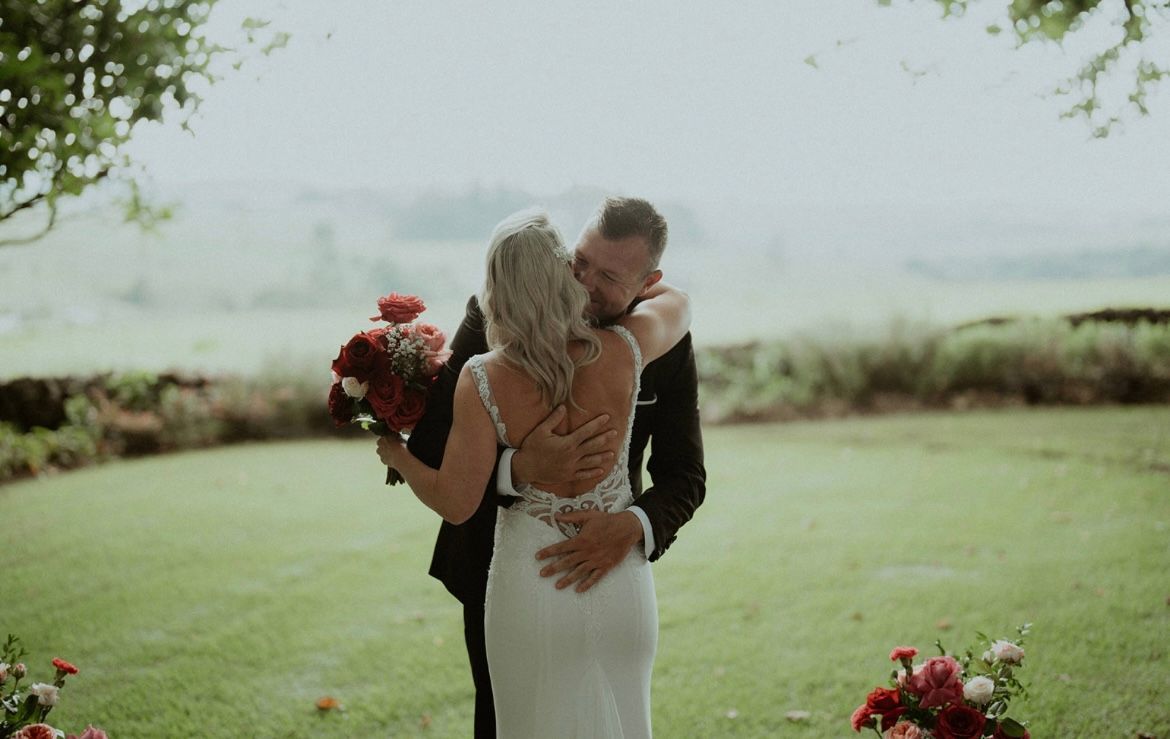 Couple embracing in a lush garden, bride holding a red bouquet during an outdoor wedding ceremony