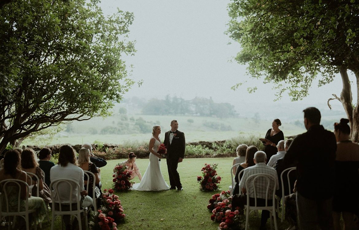 Outdoor wedding ceremony with bride and groom standing under trees, guests seated on both sides
