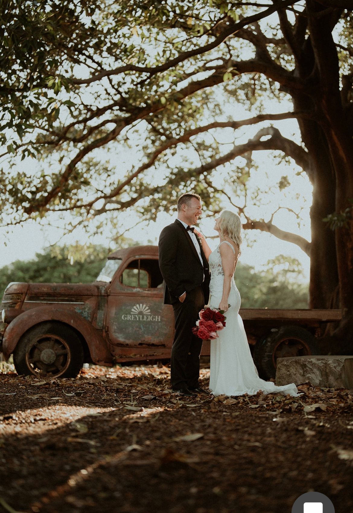 Bride and groom stand beside a vintage truck under a large tree at sunset