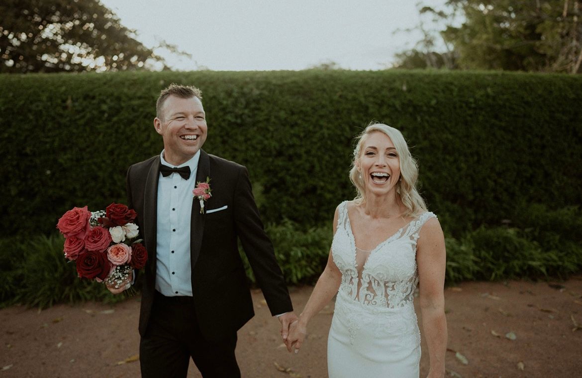 Smiling bride and groom holding hands outdoors, with flowers and a hedge backdrop.