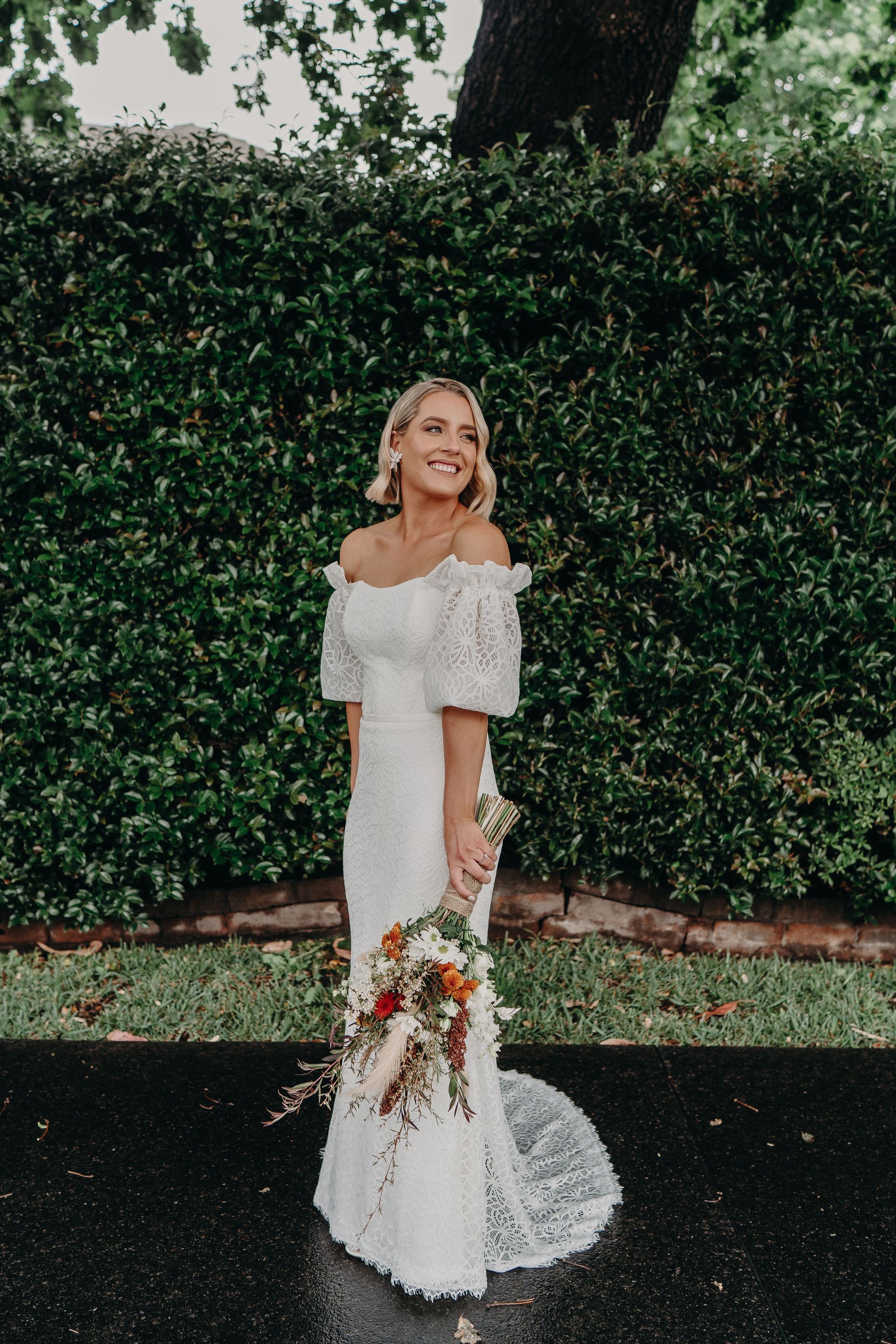 Blonde woman in white lace wedding dress smiles, holding bouquet outdoors by green hedge.