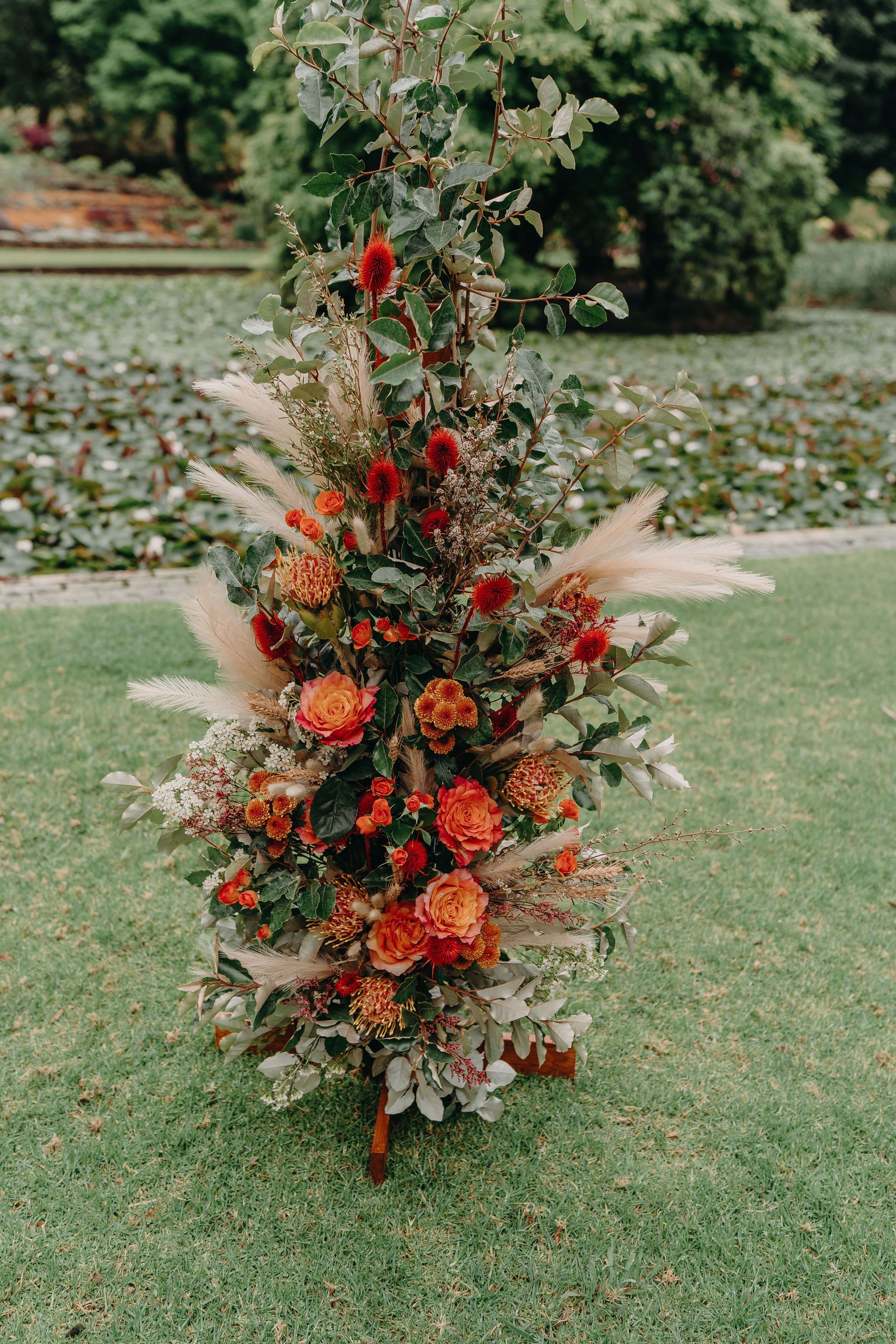 Floral arrangement with orange and red flowers, pampas grass, and foliage on green grass.