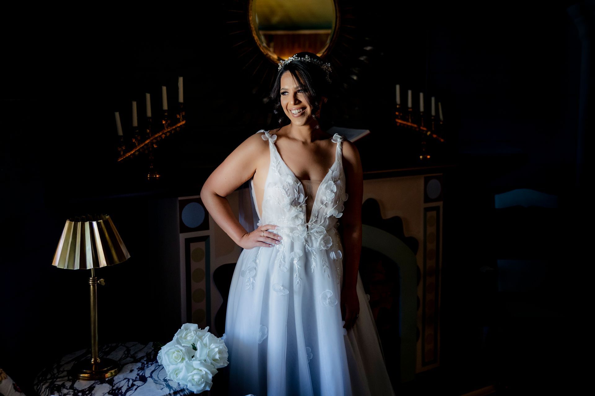 Woman in a white gown posing in a dimly lit room with a staircase and lamp in the background