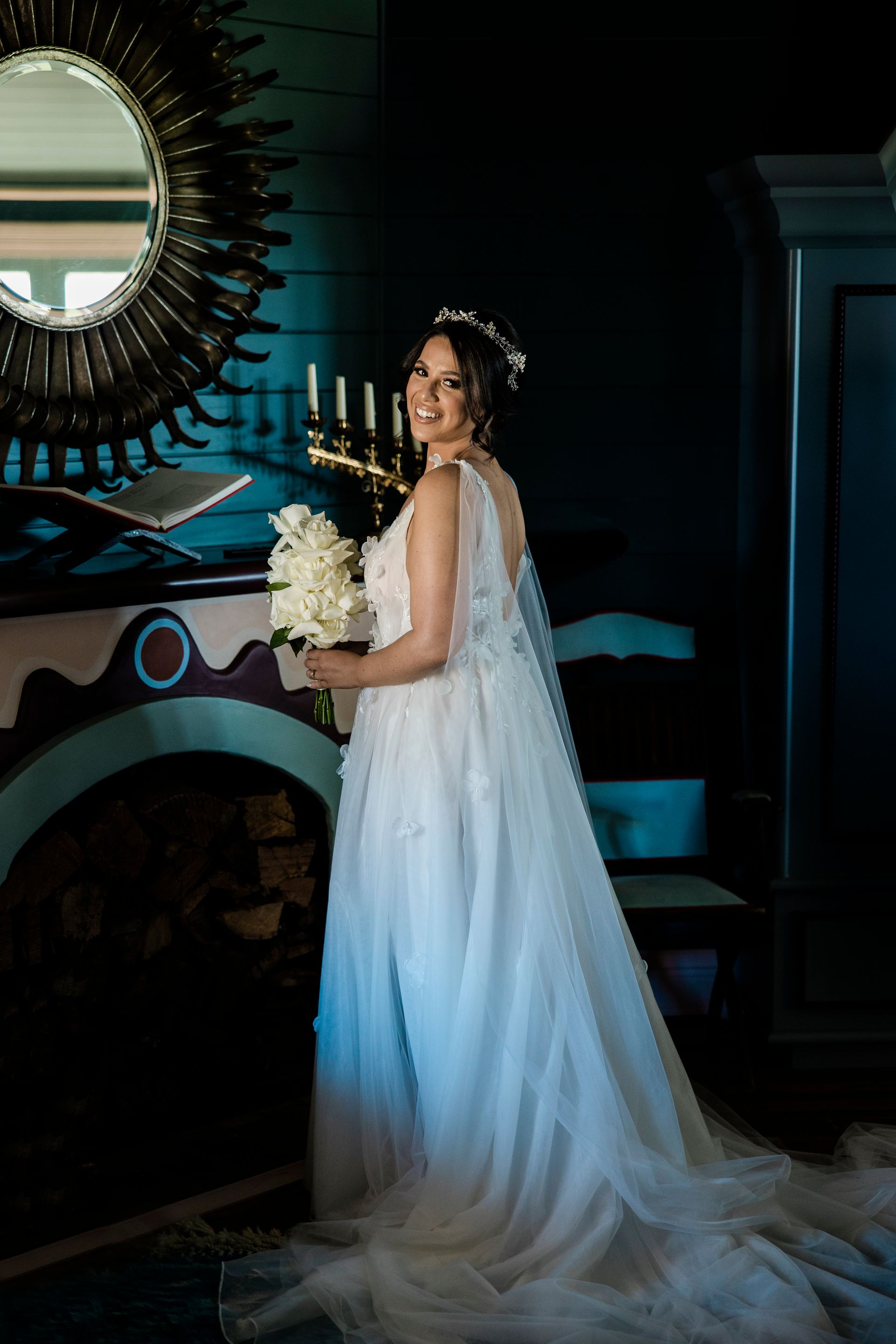 Bride in a white gown holding a bouquet, standing in a dimly lit room beside an ornate mirror.
