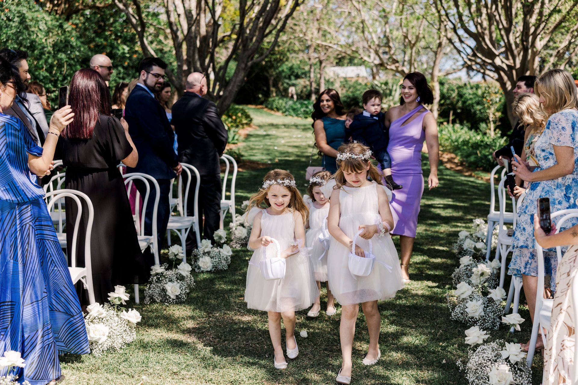 Flower girls walk down an outdoor wedding aisle between seated guests and trees.