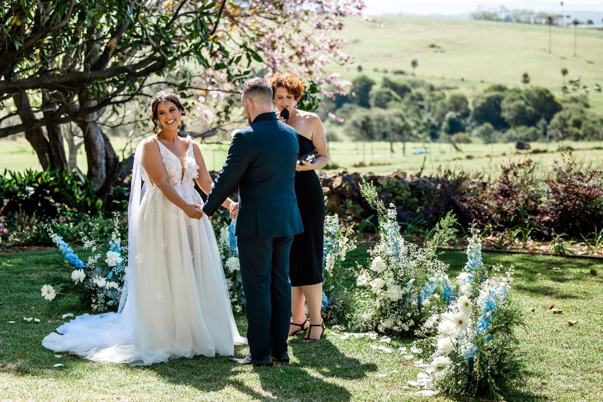 Wedding couple and officiant in a garden ceremony with white flowers and scenic hills beyond.