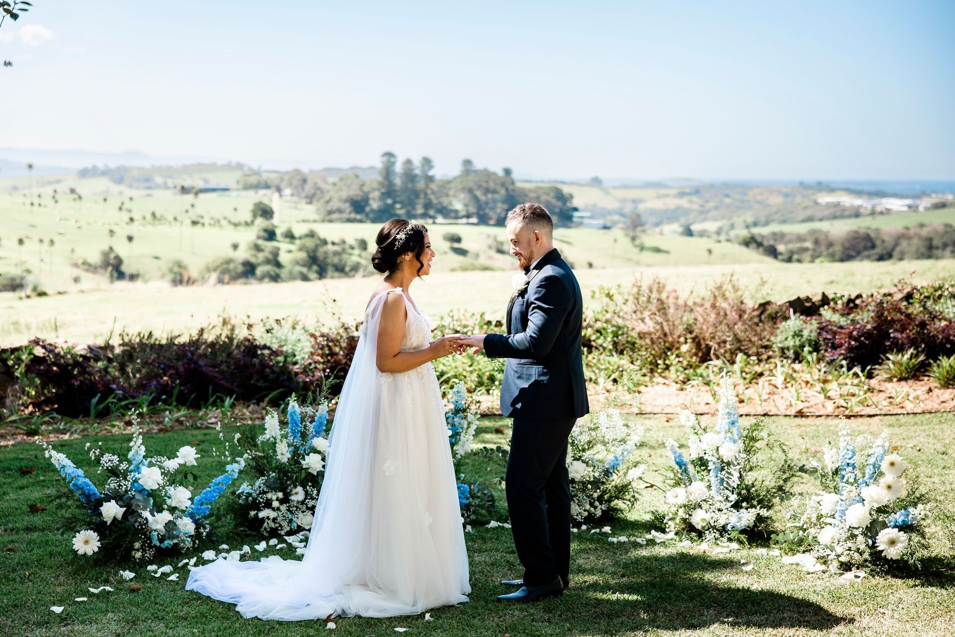 Bride and groom holding hands outdoors at a sunny wedding ceremony with floral decorations and rolling hills beyond.